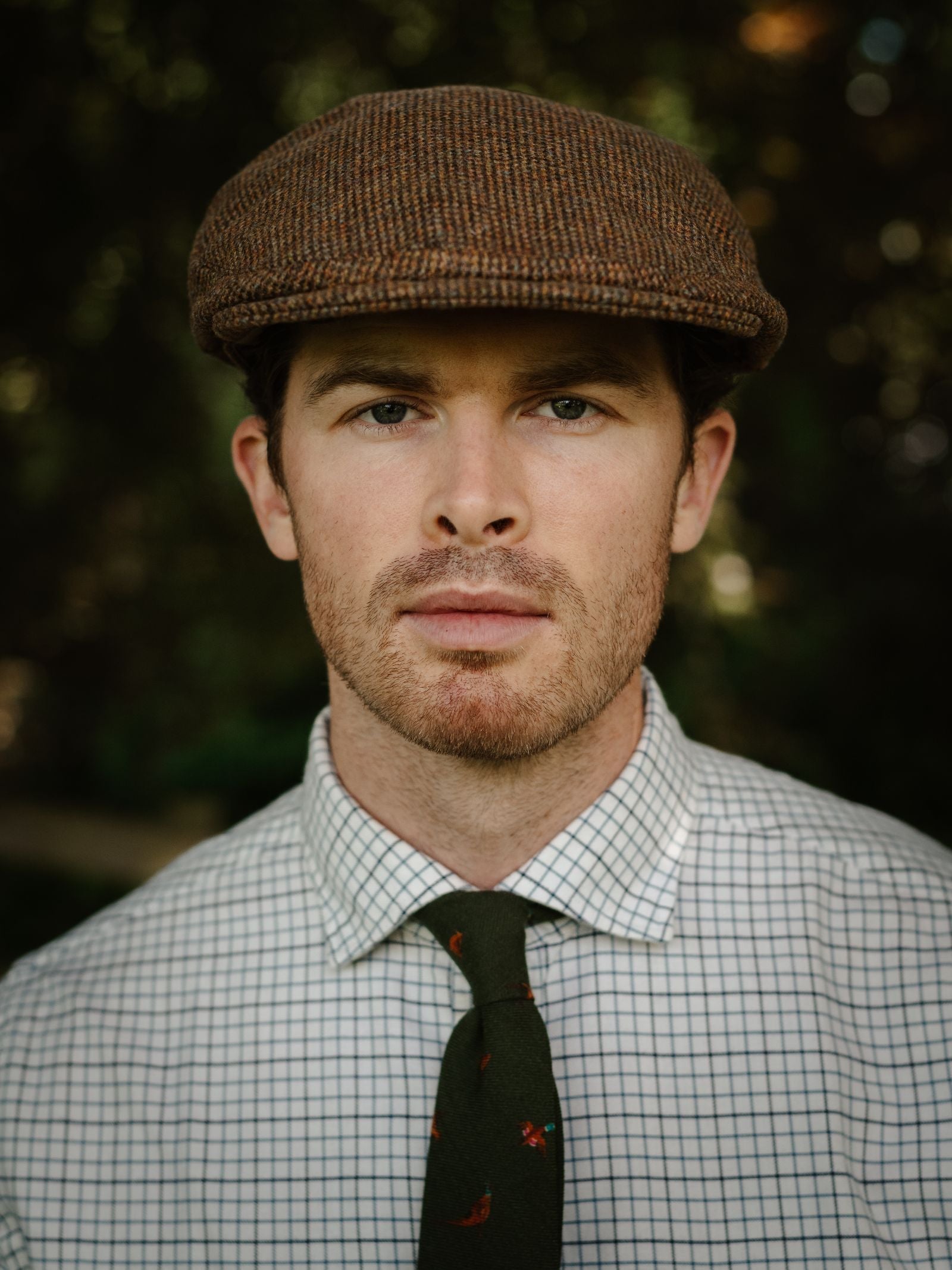 A man with light skin and short brown hair models the Unisex Beauly Cap by Campbell's of Beauly, paired with a checkered white shirt and dark green patterned tie, standing outdoors against a blurred greenery background.