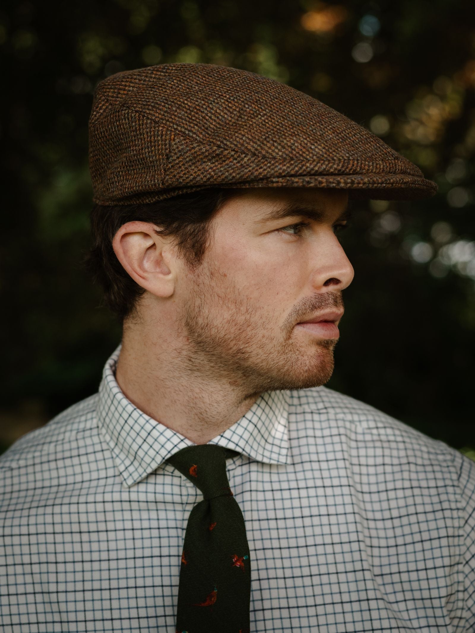 A man in the Unisex Beauly Cap by Campbell's of Beauly, paired with a white checkered shirt and dark green tie with red details, gazes to the side against a softly blurred outdoor background.