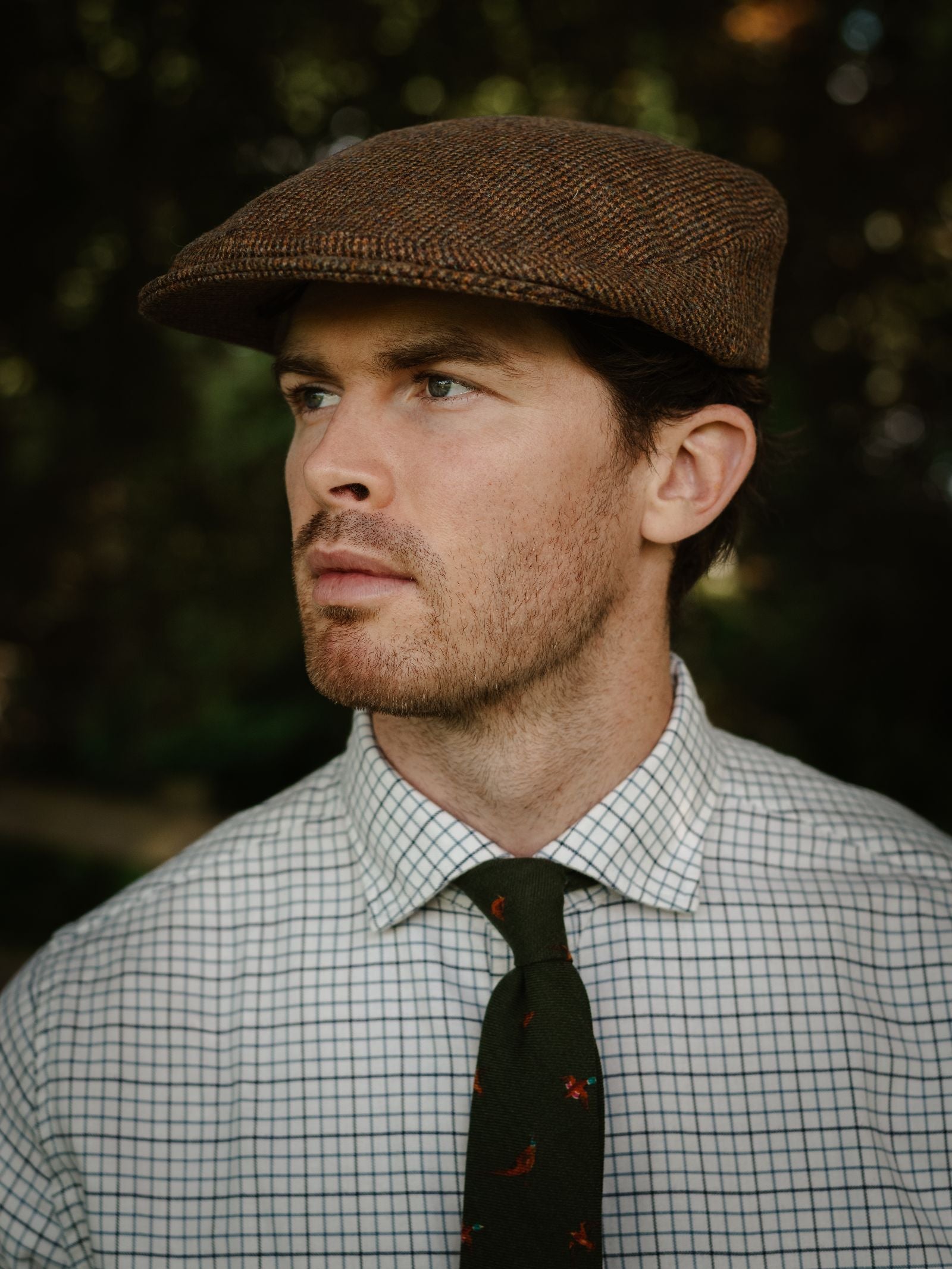 A man with light skin and short brown hair wears the Unisex Beauly Cap by Campbell's of Beauly, a white checkered shirt, and a green tie with red patterns, looking thoughtfully to the side outdoors.