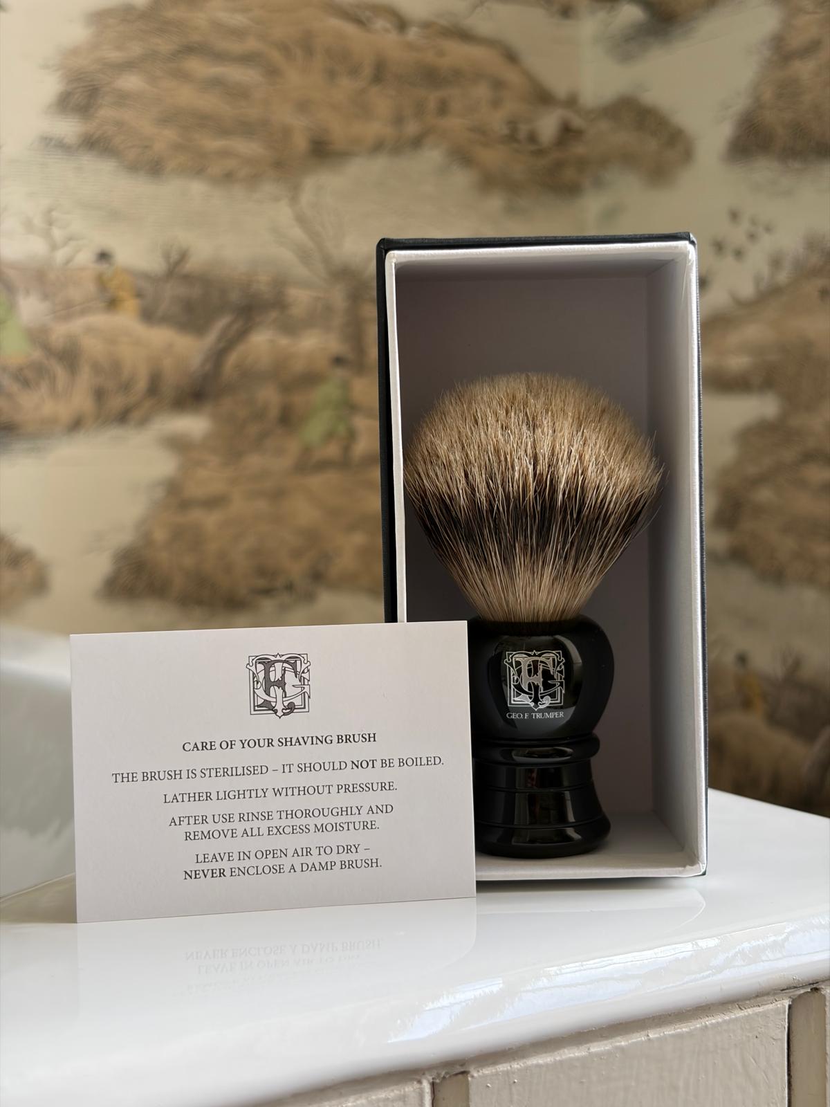 A Geo. F. Trumper Black Shaving Brush stands upright in its box with a care card in front, set on a white surface against nature-themed wallpaper—ideal for a smooth shave.