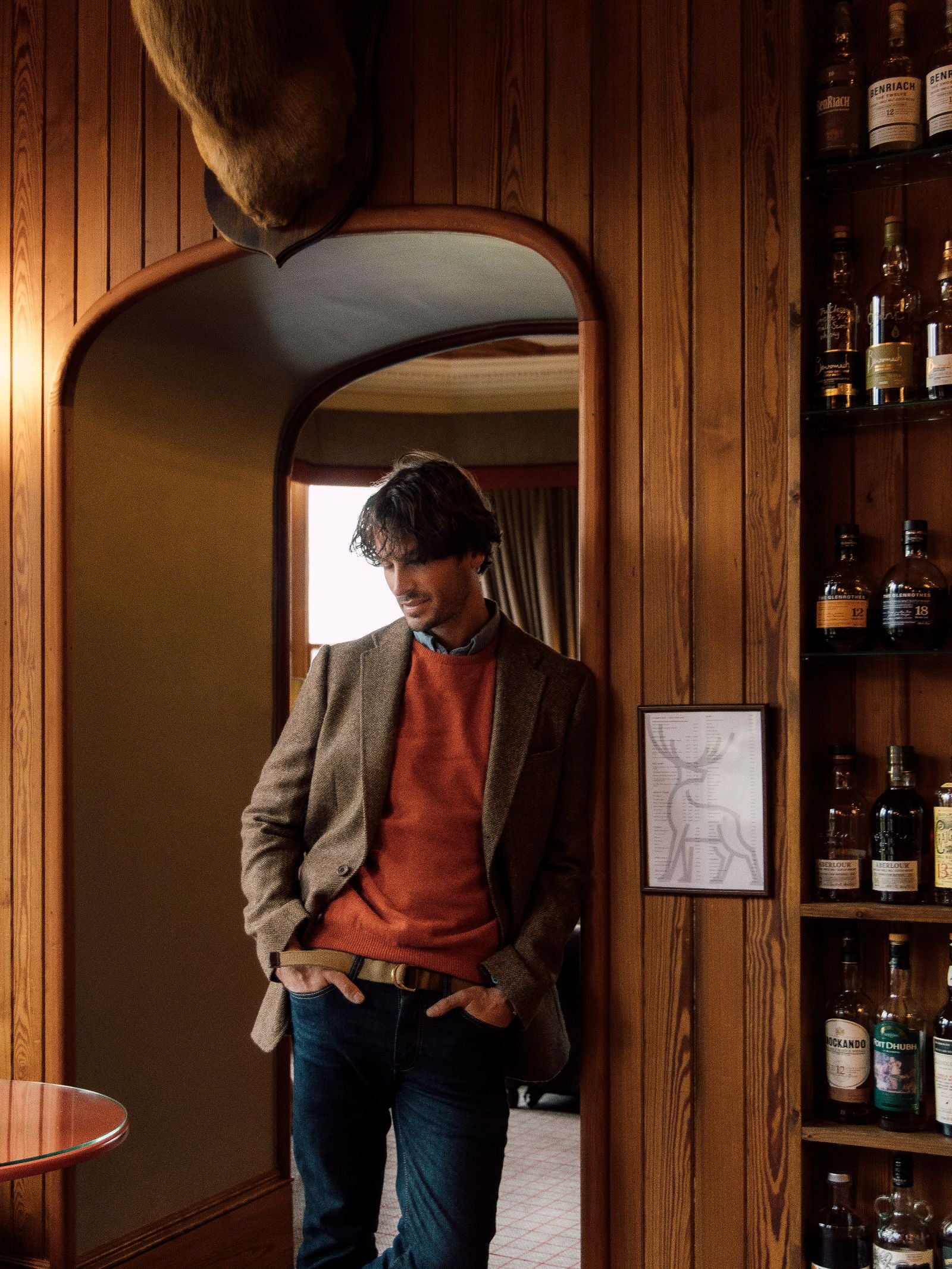 A man in a Campbell's of Beauly Campbell's House Jacket and orange sweater stands in a wood-paneled room near liquor bottles, looking down with hands in his pockets. A deer head mount hangs above the arched doorway behind him.