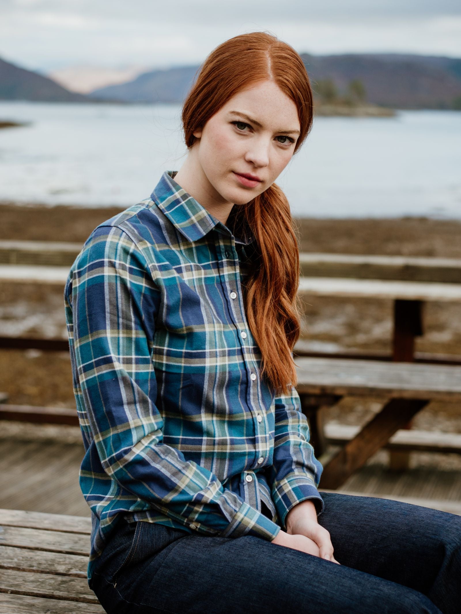 A young woman with long red hair, dressed in a Campbell's of Beauly Auld Stock Brushed Cotton Shirt and jeans, sits on a wooden bench outdoors by water and mountains, gazing calmly at the camera.
