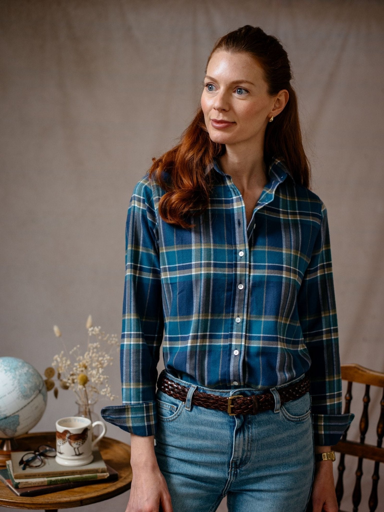 A woman with long red hair wears the Campbell's of Beauly Auld Stock Brushed Cotton Shirt and jeans, standing indoors by a wooden chair. Behind her are a table with a globe, mug, books, and dried flowers.