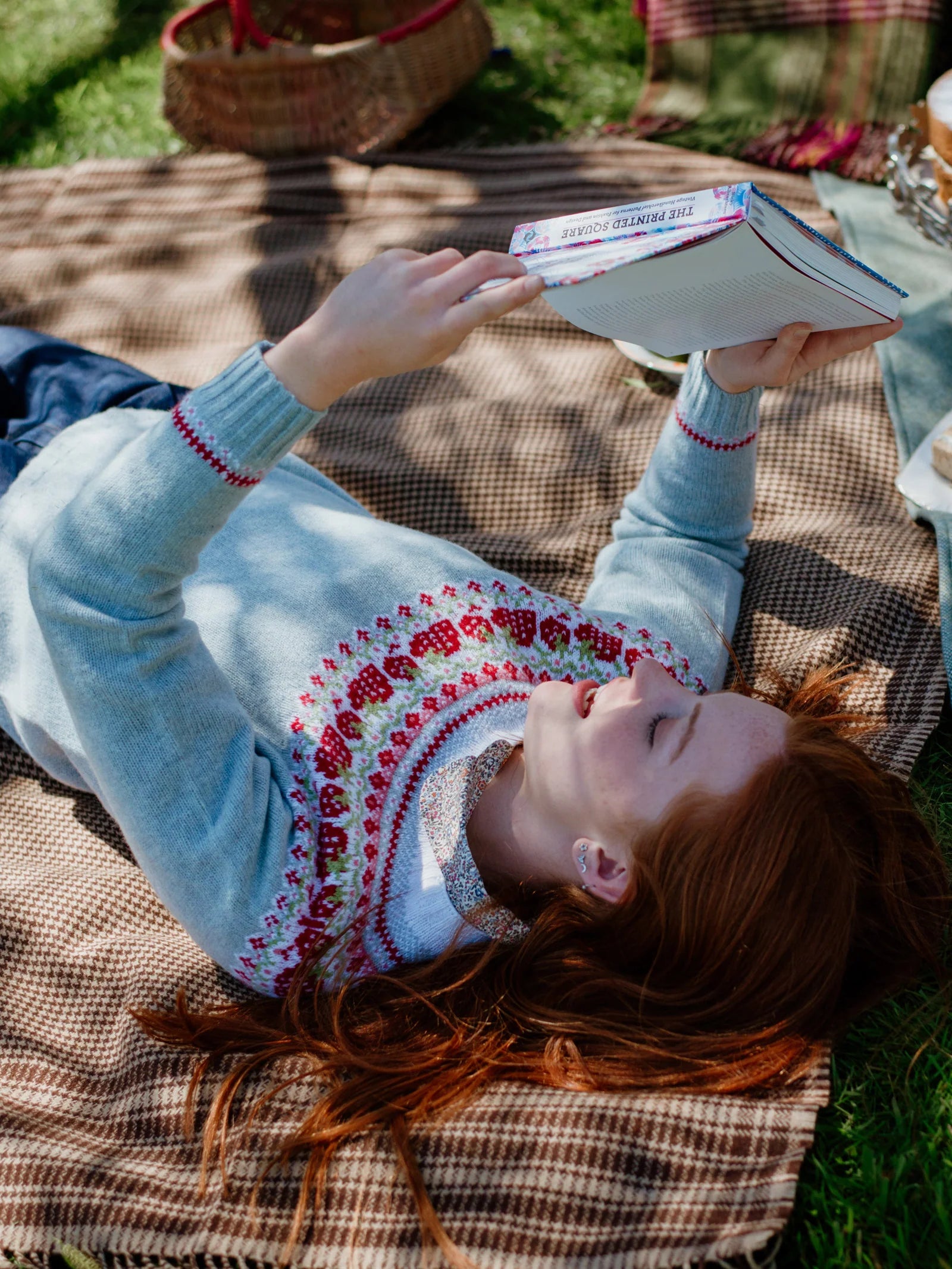 A person with long red hair relaxes outdoors on the Campbell's of Beauly Crofters Blanket, reading a book in a light blue sweater patterned in red and white, surrounded by picnic items for a cozy country vibe.