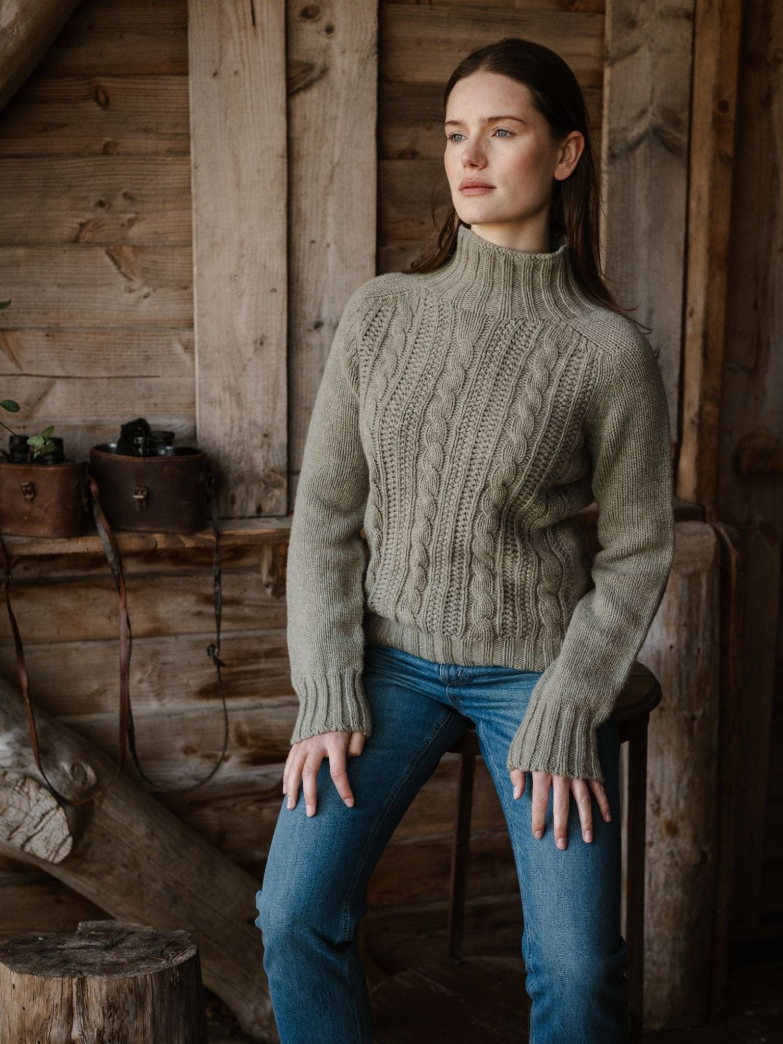 A woman in Campbell's of Beauly Cable Roll Neck Jumper, crafted in Scotland from supersoft wool, and blue jeans sits on a wooden stool in a rustic cabin, gazing thoughtfully into the distance.