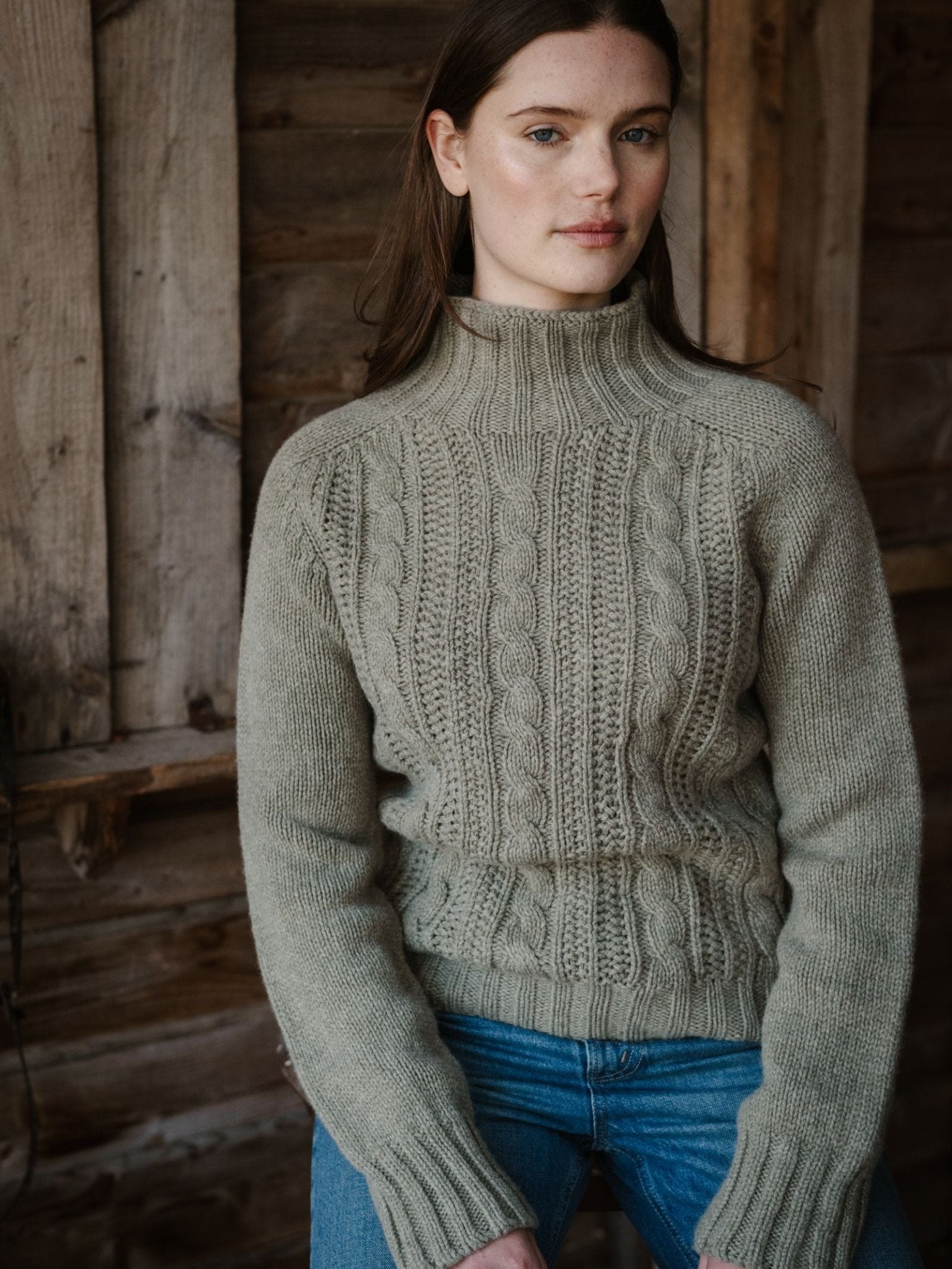 A woman with long brown hair wearing the Campbell's of Beauly Cable Roll Neck Jumper in light beige and blue jeans sits by a rustic wooden wall, looking calmly at the camera. This supersoft wool sweater is beautifully knitted in Scotland.