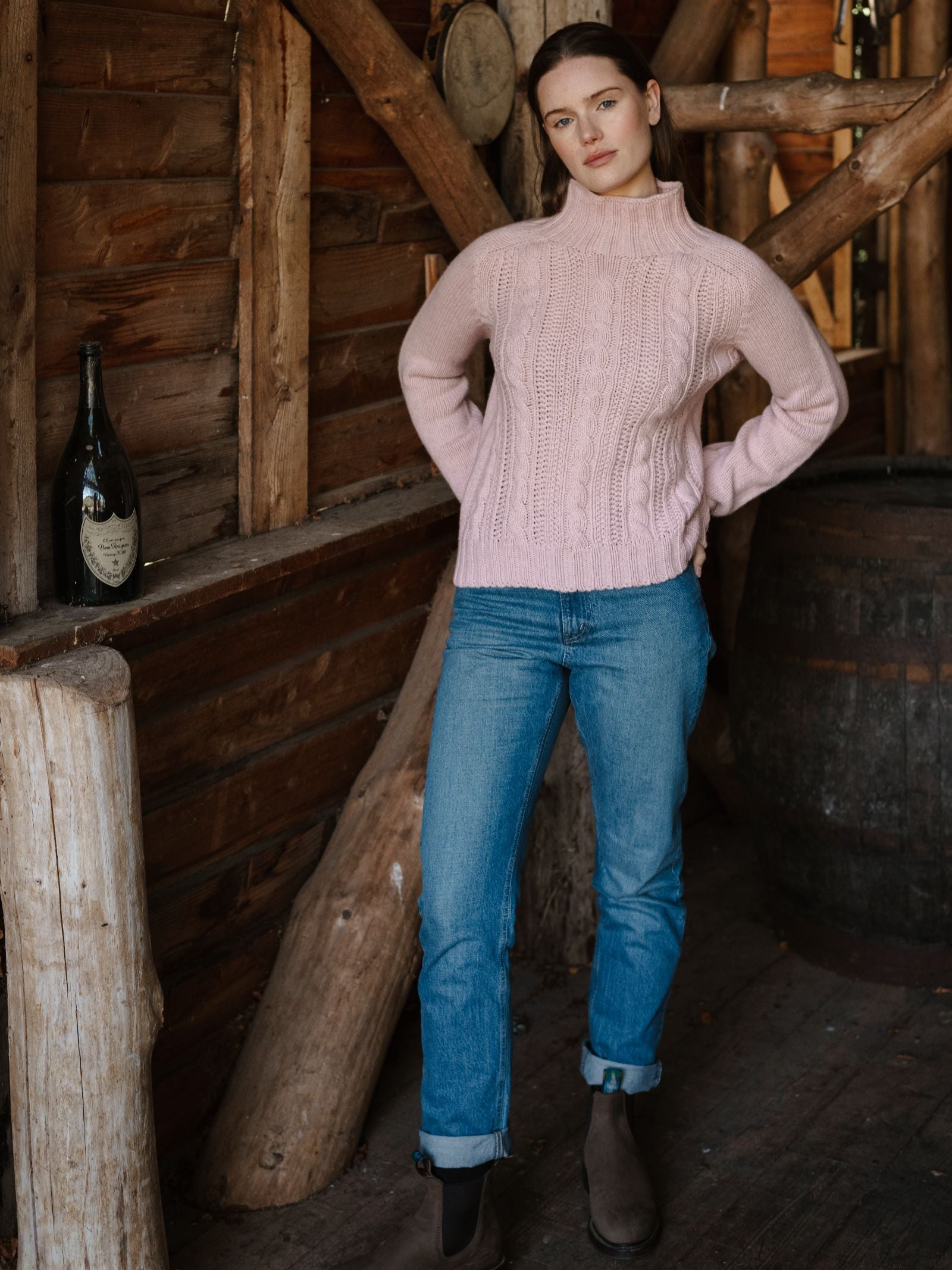 A woman wears the Campbell's of Beauly Cable Roll Neck Jumper, knitted in Scotland from supersoft wool, with blue jeans and boots. She stands indoors against a rustic wooden wall with a large wine bottle and barrel behind her.