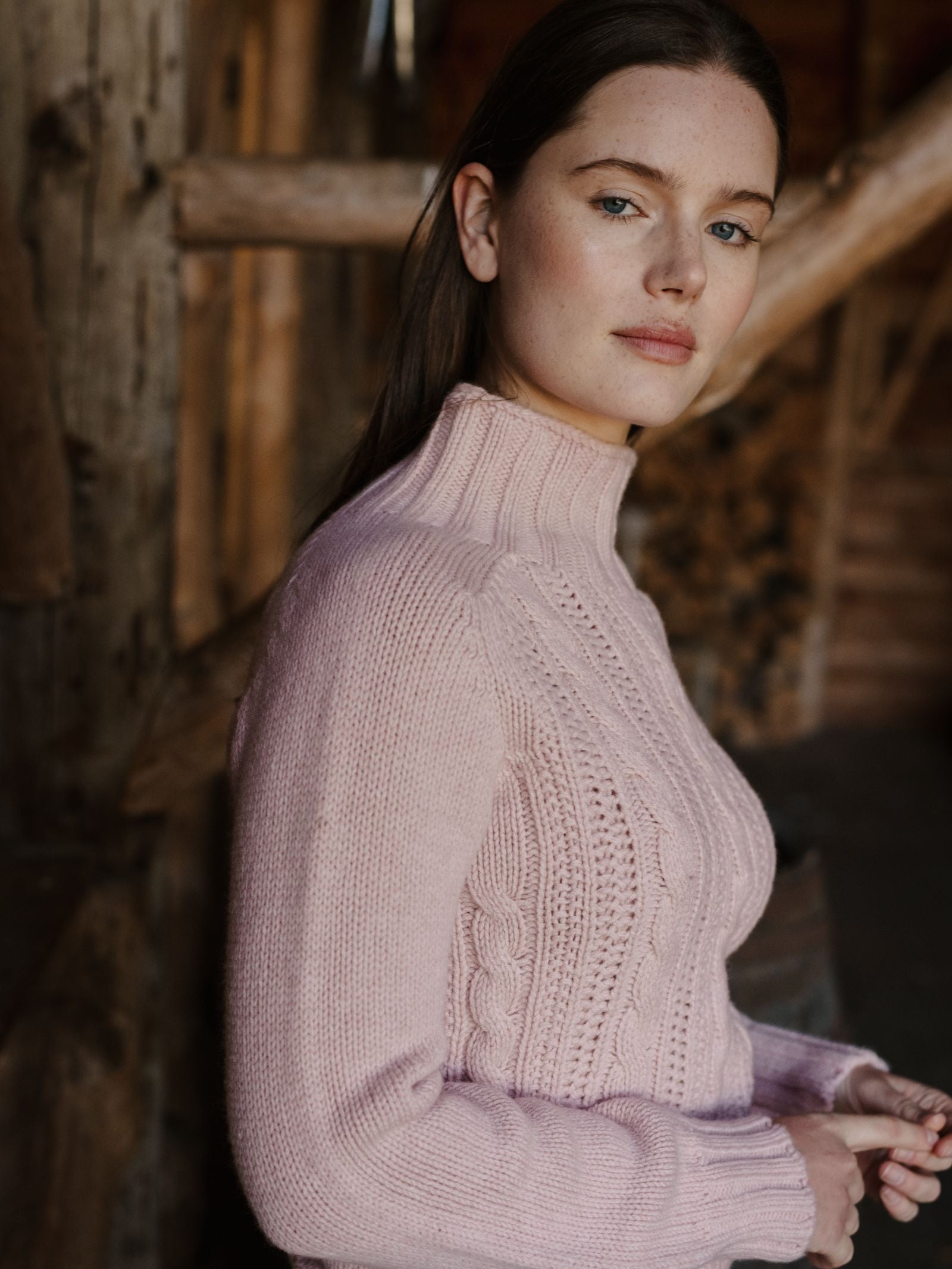 A woman with long brown hair wears the Campbell's of Beauly Cable Roll Neck Jumper in Rose, a supersoft wool sweater knitted in Scotland, standing indoors before rustic wooden beams and looking calmly at the camera.