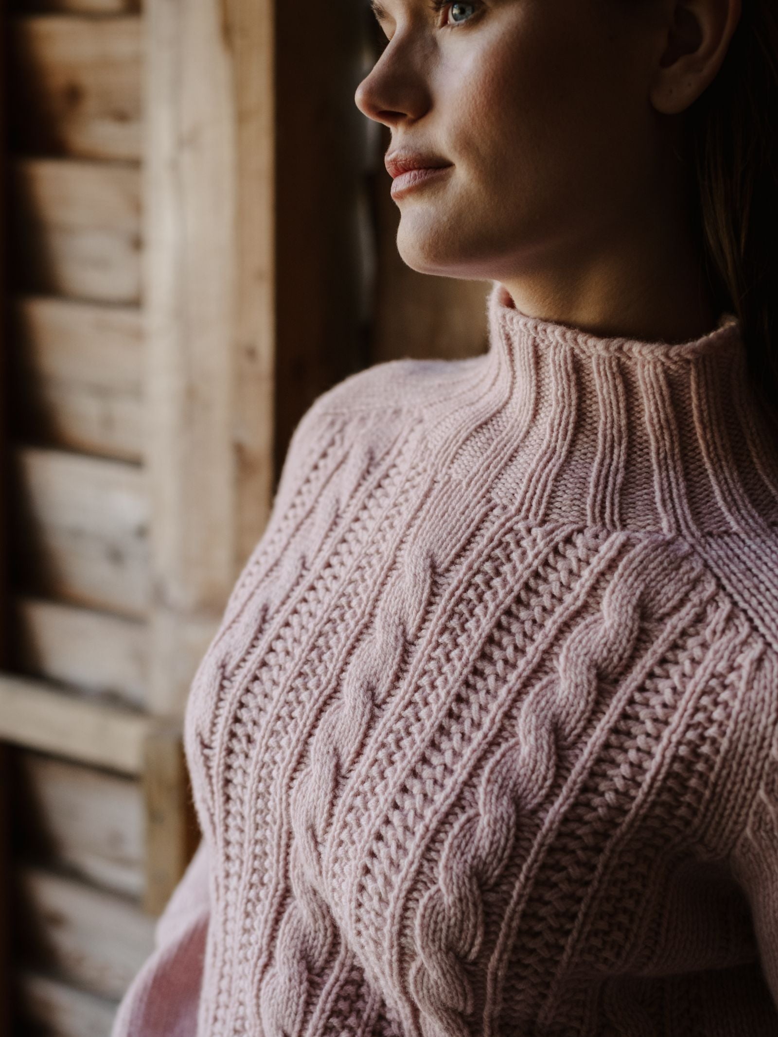 A woman stands indoors by a wooden wall, natural light highlighting her Cable Roll Neck Jumper by Campbell’s of Beauly—a light pink, thick cable-knit sweater crafted in Scotland from supersoft wool yarns.