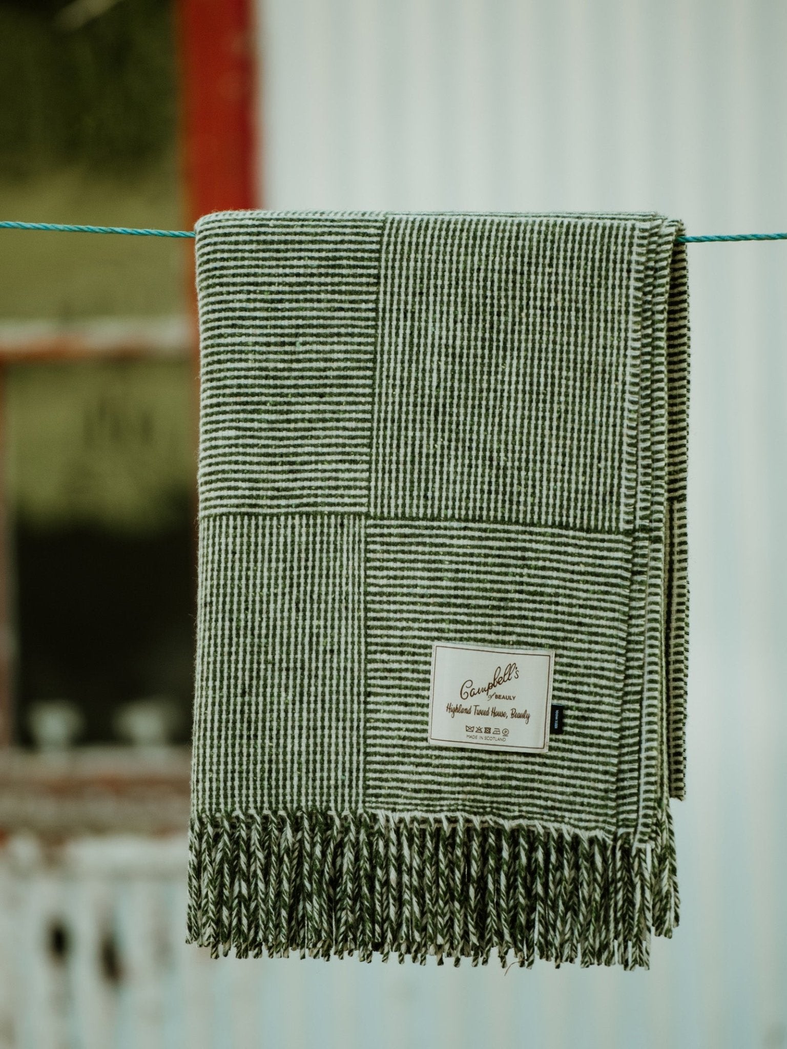The Cabrich Lambswool Throw by Campbells of Beauly, featuring green and white stripes with fringe, hangs over a blue clothesline. Its label is visible; a blurred window and corrugated wall appear in the background.