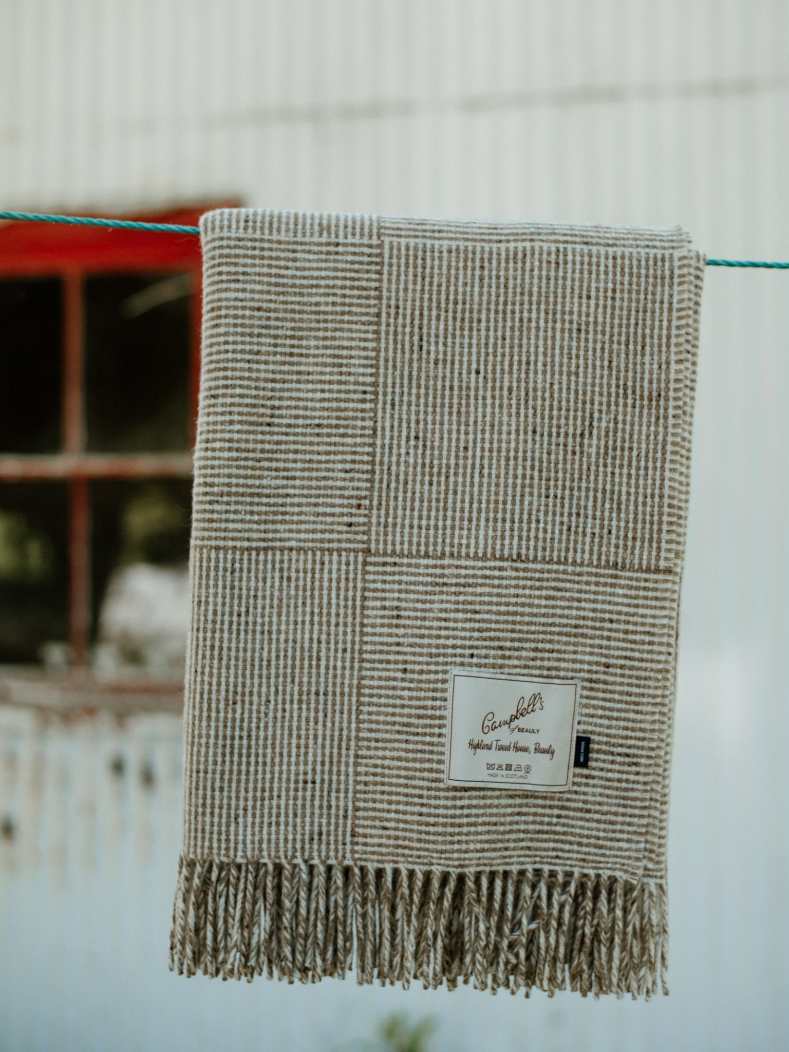 The Cabrich Lambswool Throw by Campbells of Beauly, featuring a beige and brown patchwork basket weave and fringe, hangs outdoors on a line. A visible label accents the blanket; a blurred window and corrugated wall are behind.
