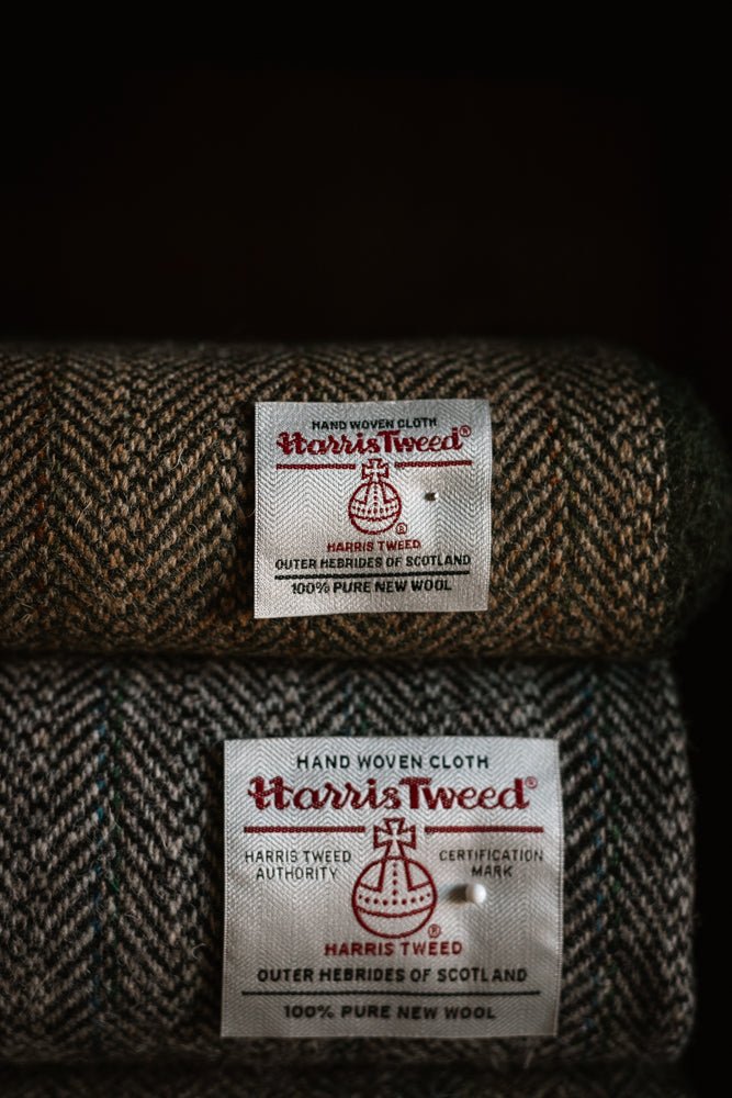 Two rolls of House Tweed 95054 by Campbell’s of Beauly, each featuring brown and green patterned fabric with a white label bearing the Harris Tweed Authority orb logo and certifying it is handwoven in Scotland from 100% pure new wool. These distinguished jacketing cloths boast a classic herringbone design, perfect for timeless elegance.