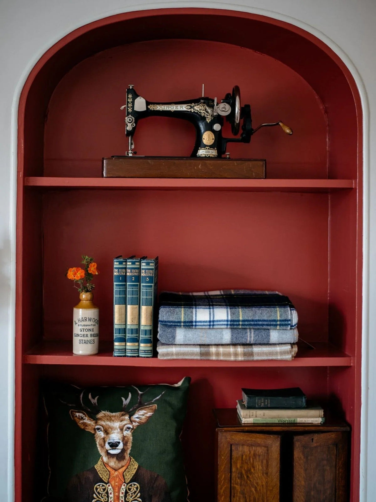 A vintage sewing machine, books, a jar with flowers, folded Campbell’s of Beauly Lambswool Tartan Blankets, a pillow featuring a deer in a suit, and two small books are displayed on red shelves built into a wall alcove—blending heritage charm with UK made accents.
