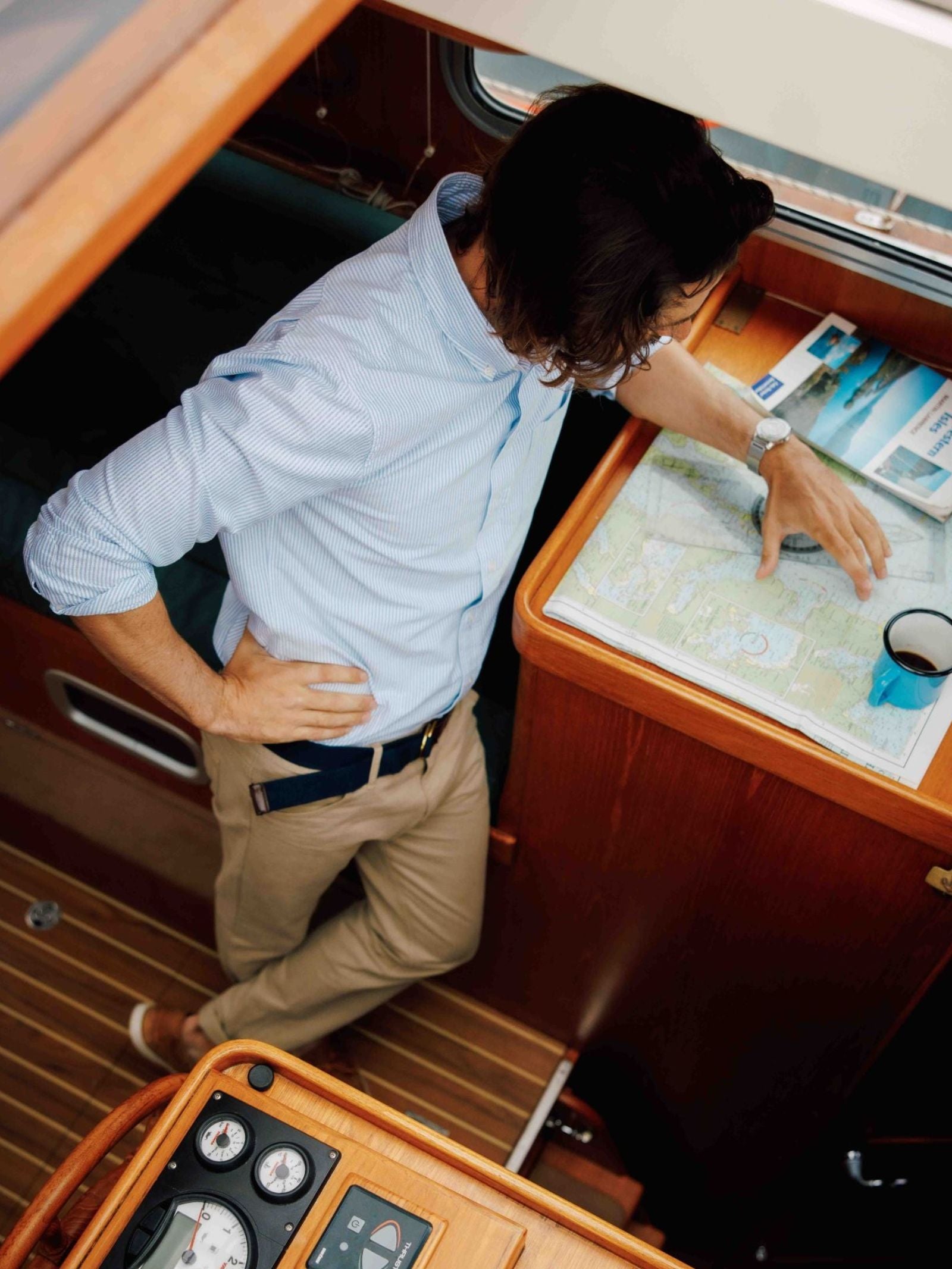 A person stands inside a wooden boat cabin, looking at a map on a table with a coffee cup nearby. The person wears a blue shirt, khaki pants, and the Arnold Wills & Co Ltd Canvas D-Loop Belt in navy, which adds to their country wardrobe, while navigation instruments are visible in the foreground.
