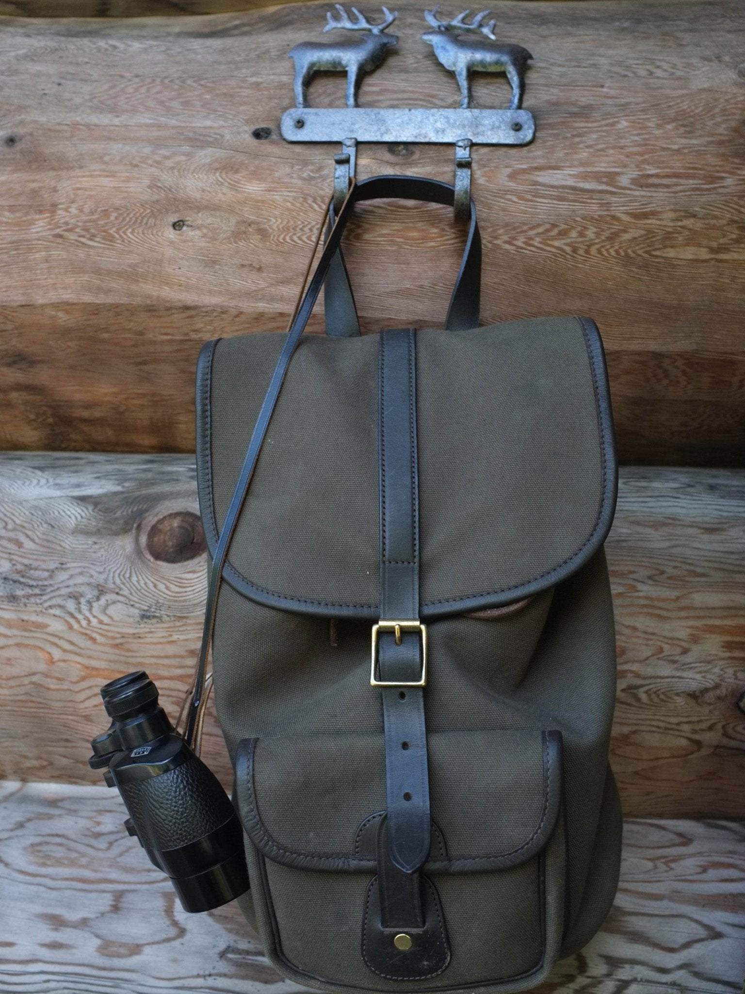 The Campbell's of Beauly Canvas Rucksack with brown leather trim hangs on a rustic wooden wall from a deer-shaped metal hook, with binoculars attached to the side.