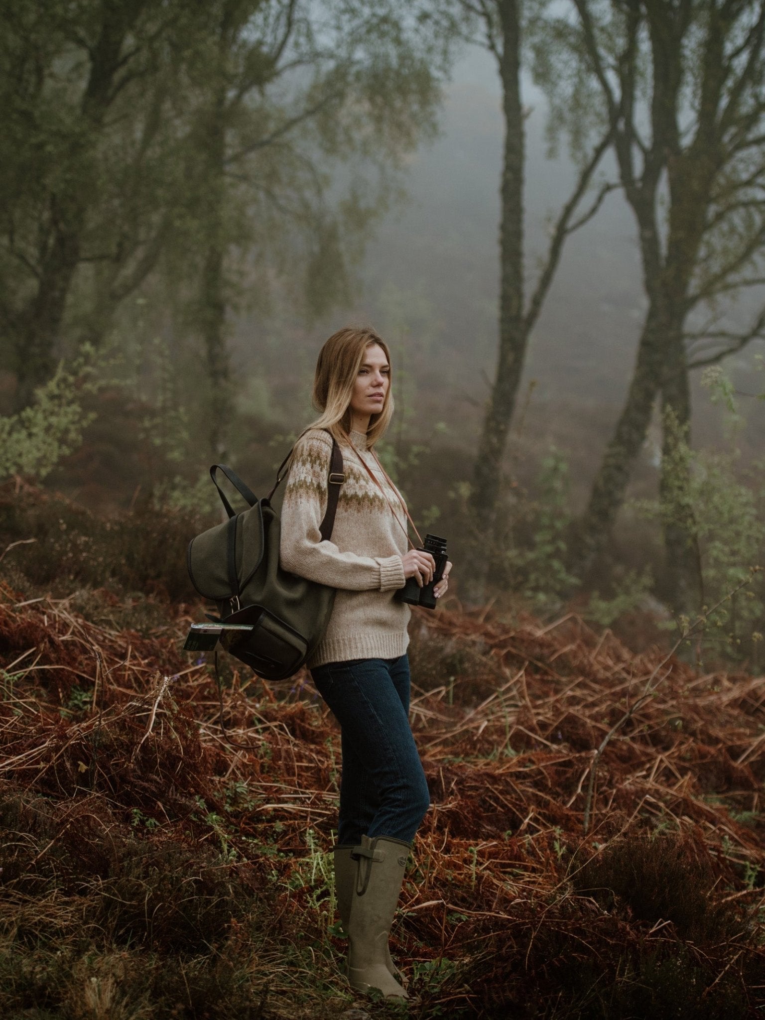 A woman stands in a foggy forest, surrounded by brown ferns and mossy trees, holding binoculars and a camera. She carries the Campbell's of Beauly Canvas Rucksack, a green canvas bag with leather trim.