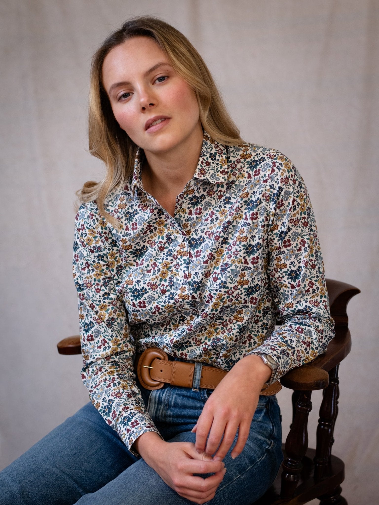 A woman with long blonde hair sits on a wooden chair, wearing a floral button-up shirt, blue jeans, and the Caramel Leather Belt by Campbell's of Beauly, posing against a neutral fabric background.
