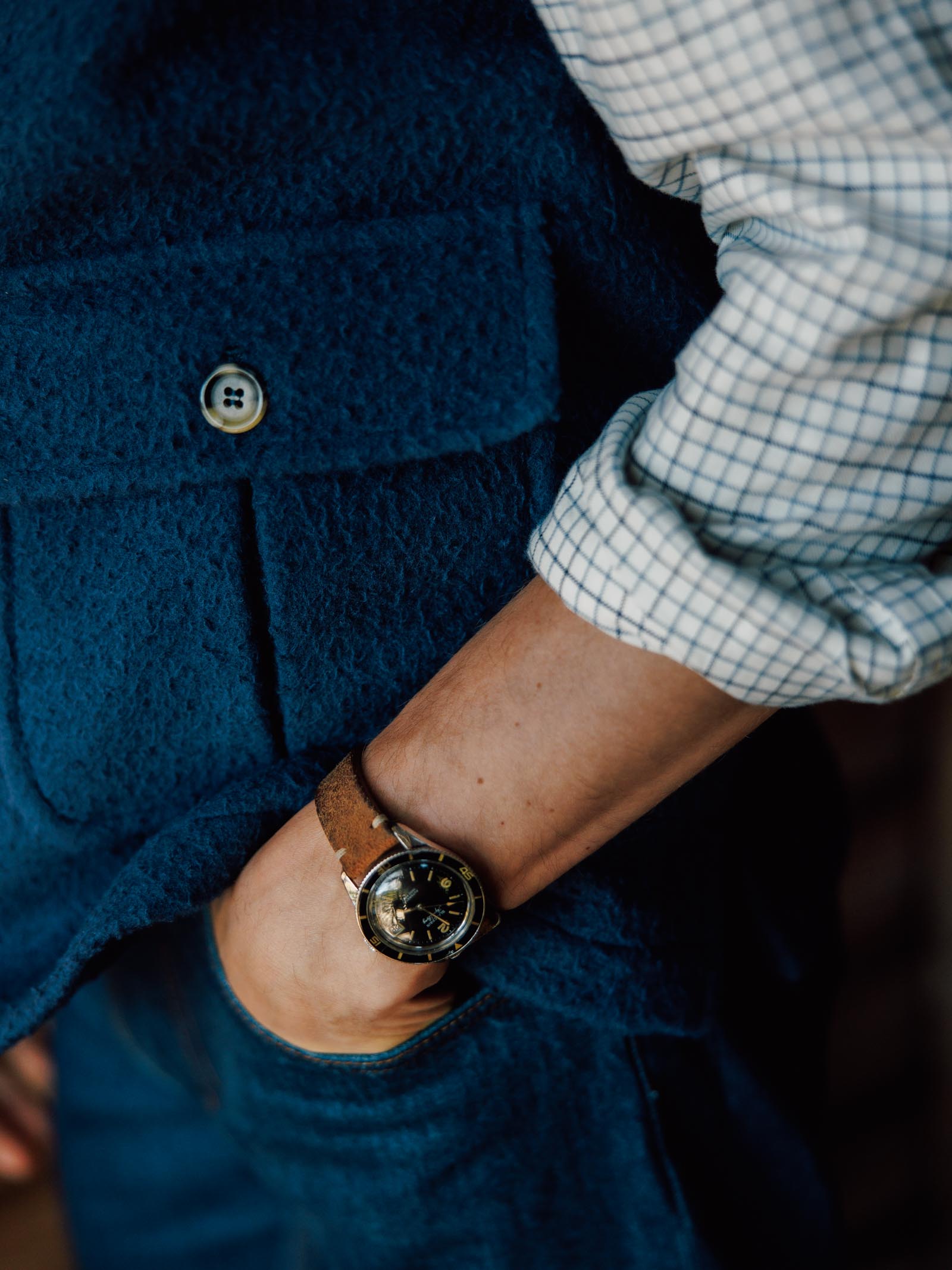 A person in a blue Campbell's of Beauly Casentino Fleece Gilet and checkered shirt stands with one hand in their jeans pocket, highlighting a brown leather wristwatch and the outfit's classic country style details.