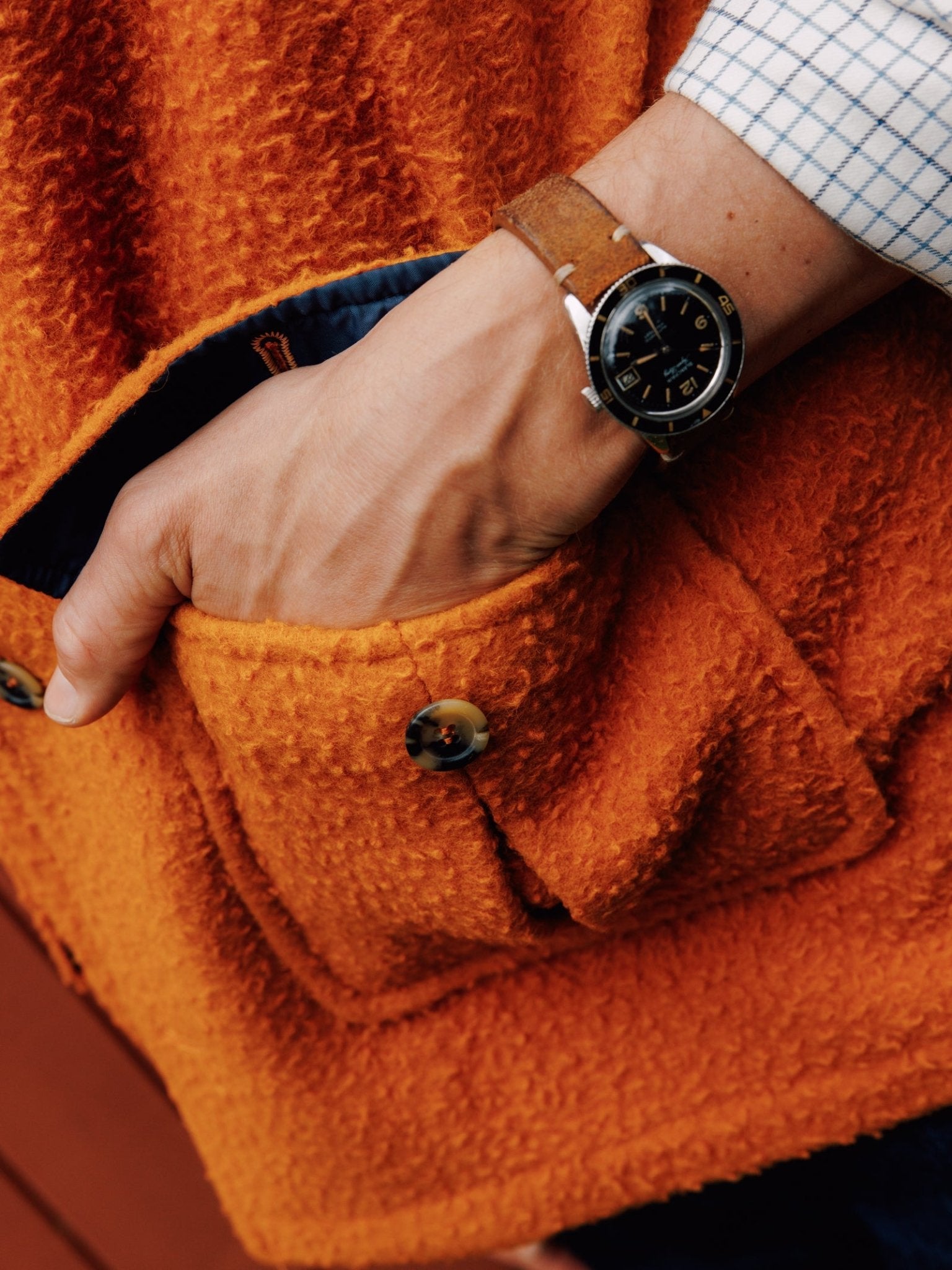 A person in a blue checkered shirt with a brown wristwatch has their hand in the pocket of an orange Casentino Fleece Gilet by Campbell's of Beauly, which features brown buttons.