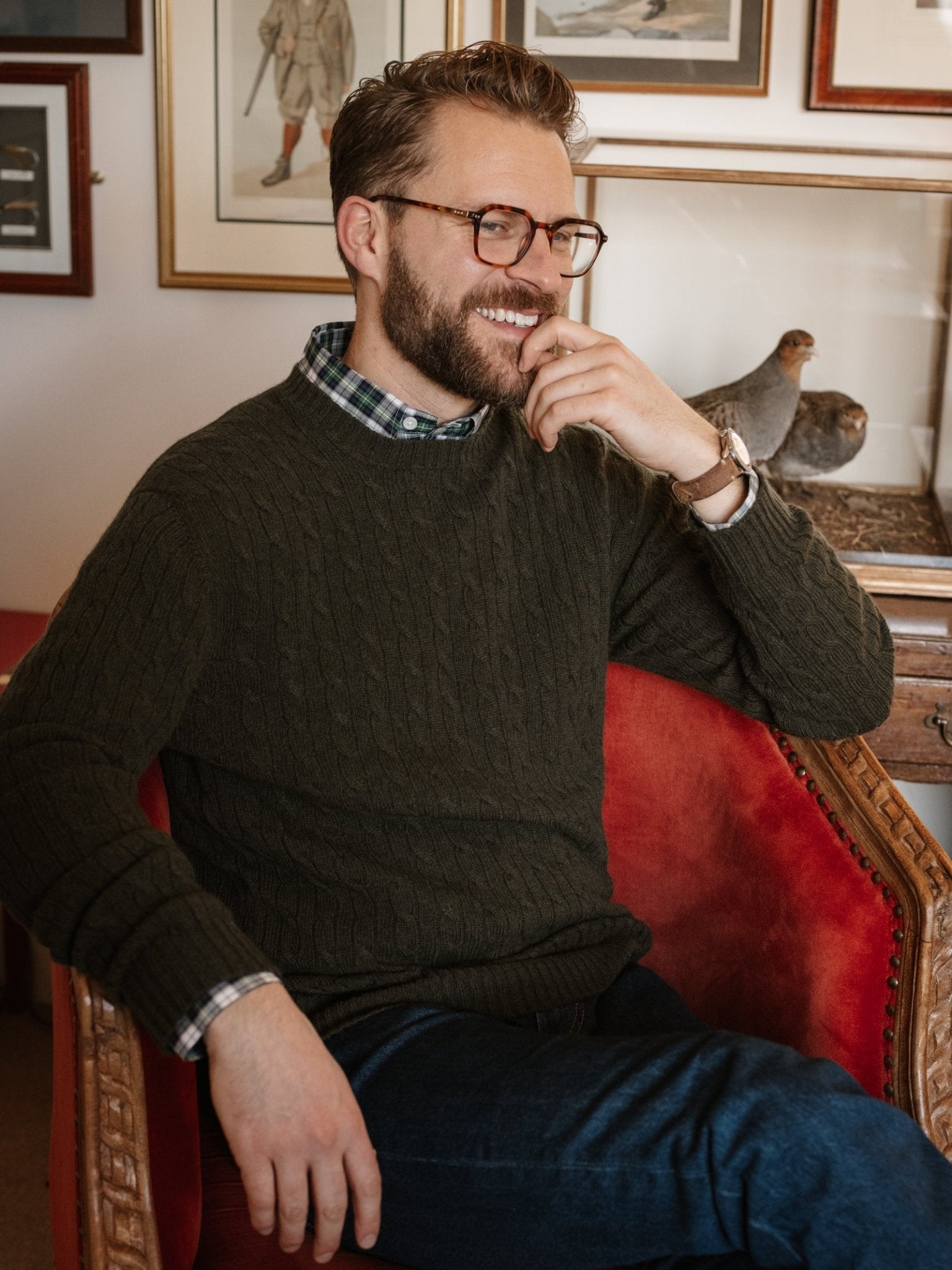 A bearded man with glasses smiles while wearing a green Campbell's of Beauly Cashmere Cable Crew Jumper over a plaid shirt, seated in a red armchair. Framed artwork and bird figurines decorate the background.