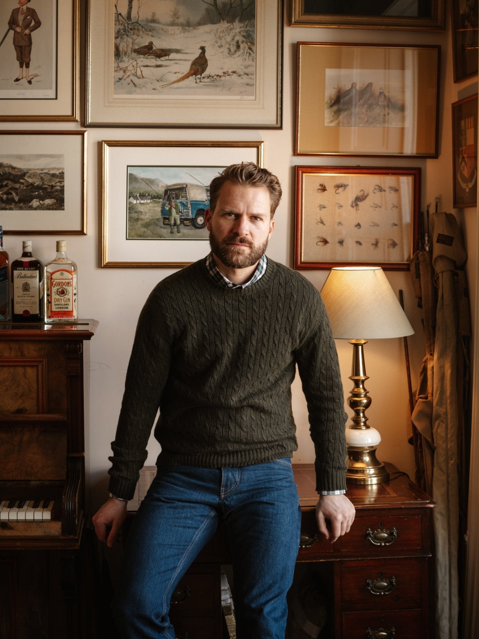 A bearded man in a Campbell's of Beauly Cashmere Cable Crew Jumper and blue jeans sits on a wooden desk beside a lamp, with framed artwork and vintage paintings decorating the wall behind him.