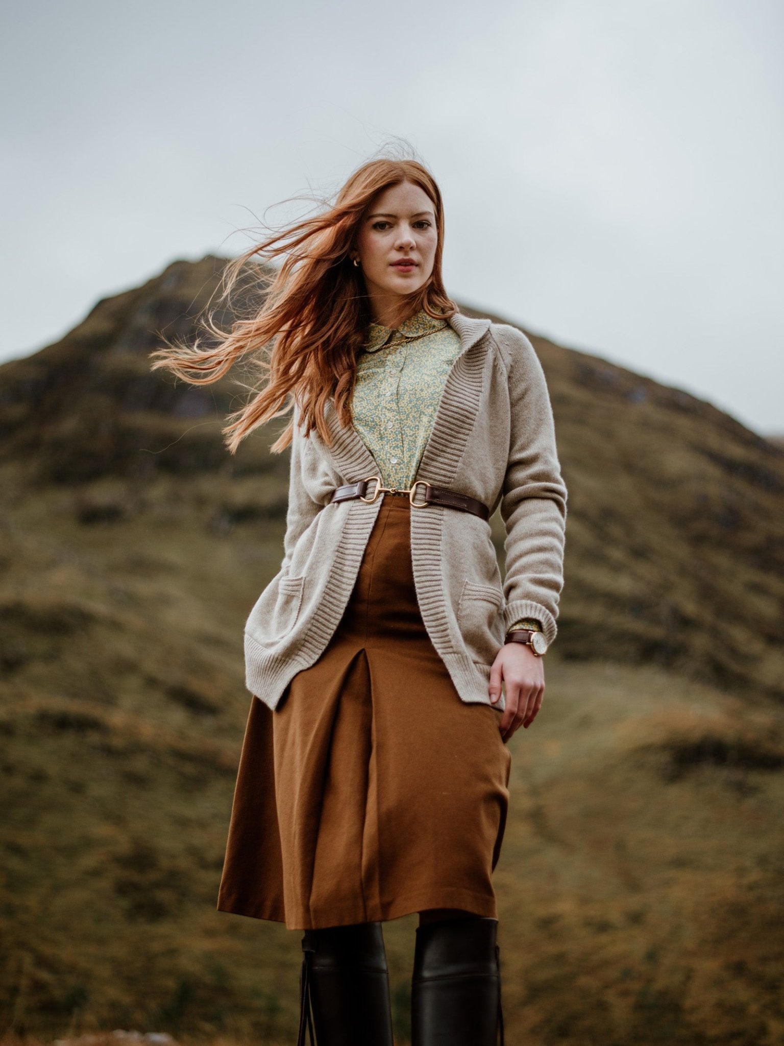 A woman with long red hair stands outdoors in front of a grassy hill, wearing the Campbell's of Beauly Cashmere Shawl Cardigan, a patterned blouse, brown skirt, and black boots. Her windblown hair and confident gaze capture the moment.