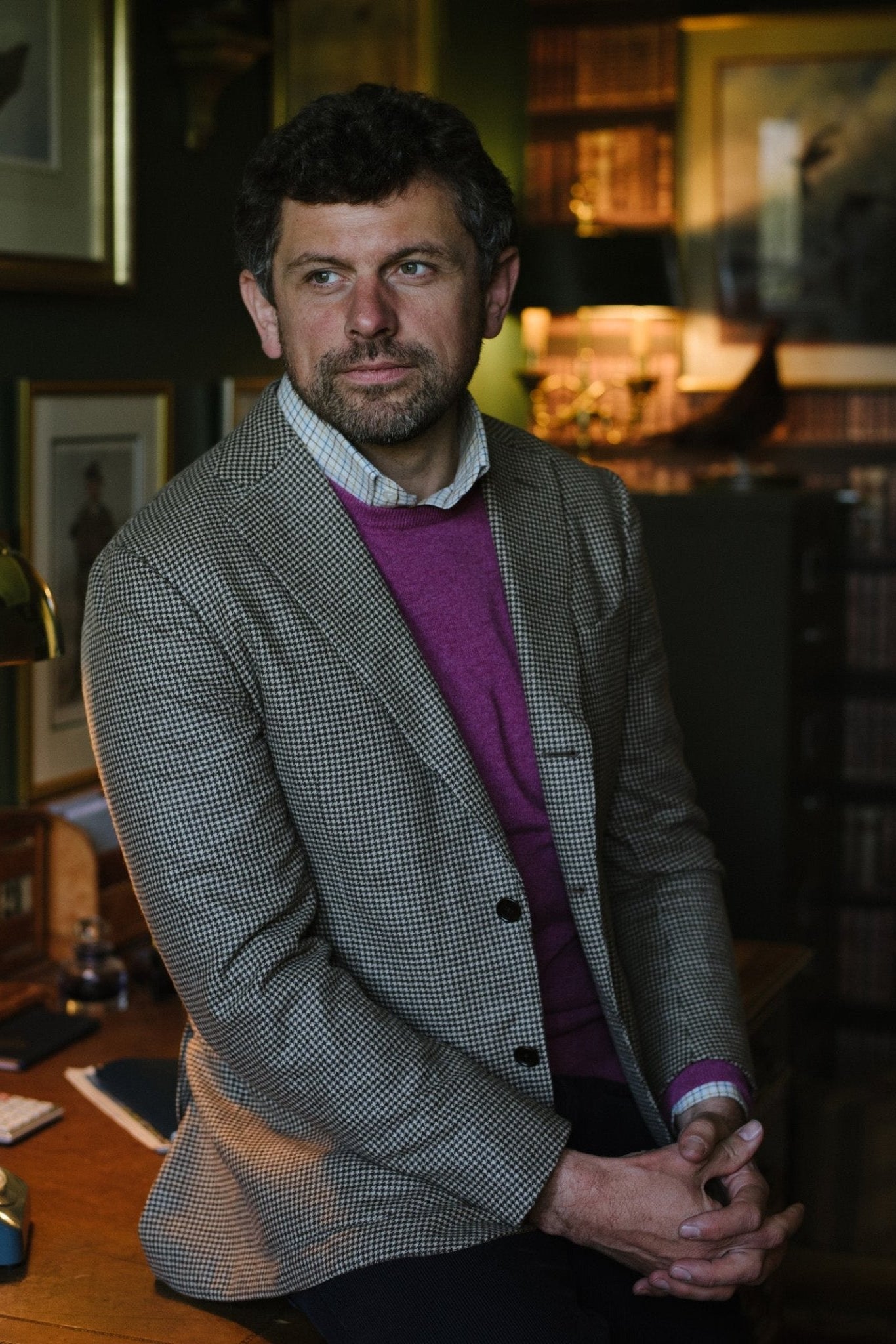 A man with short curly hair and a beard, wearing a gray checked blazer over the Campbell's of Beauly Auld Stock Cashmere Crew Neck Jumper, sits on a wooden desk in a warmly lit study with bookshelves and framed pictures behind him.