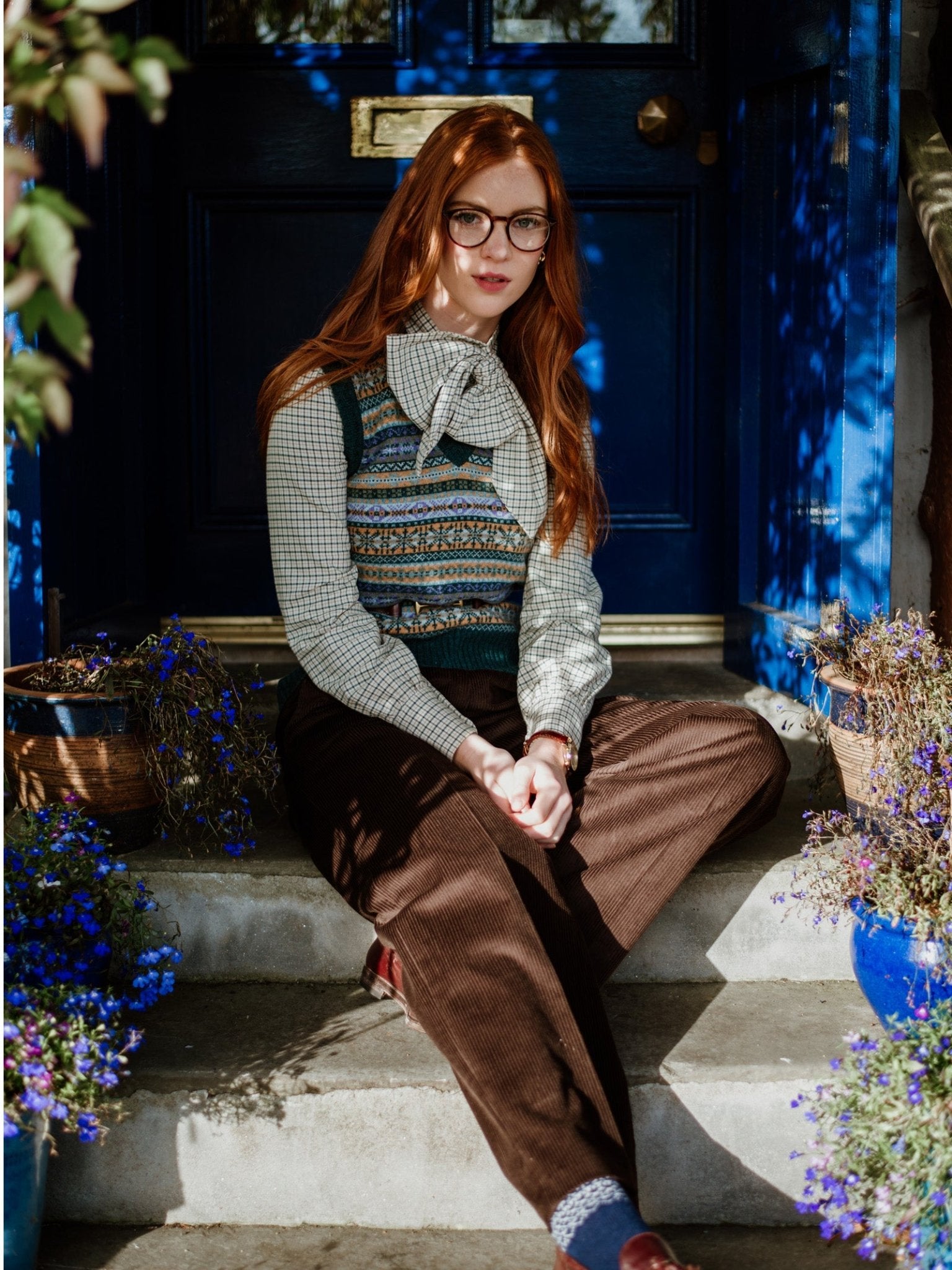 A woman with long red hair and glasses sits on outdoor steps by a blue door, wearing the Campbell's of Beauly Auld Stock Cashmere V-Neck Fairisle Slipover over a checked shirt, paired with brown corduroy pants and surrounded by purple potted flowers.