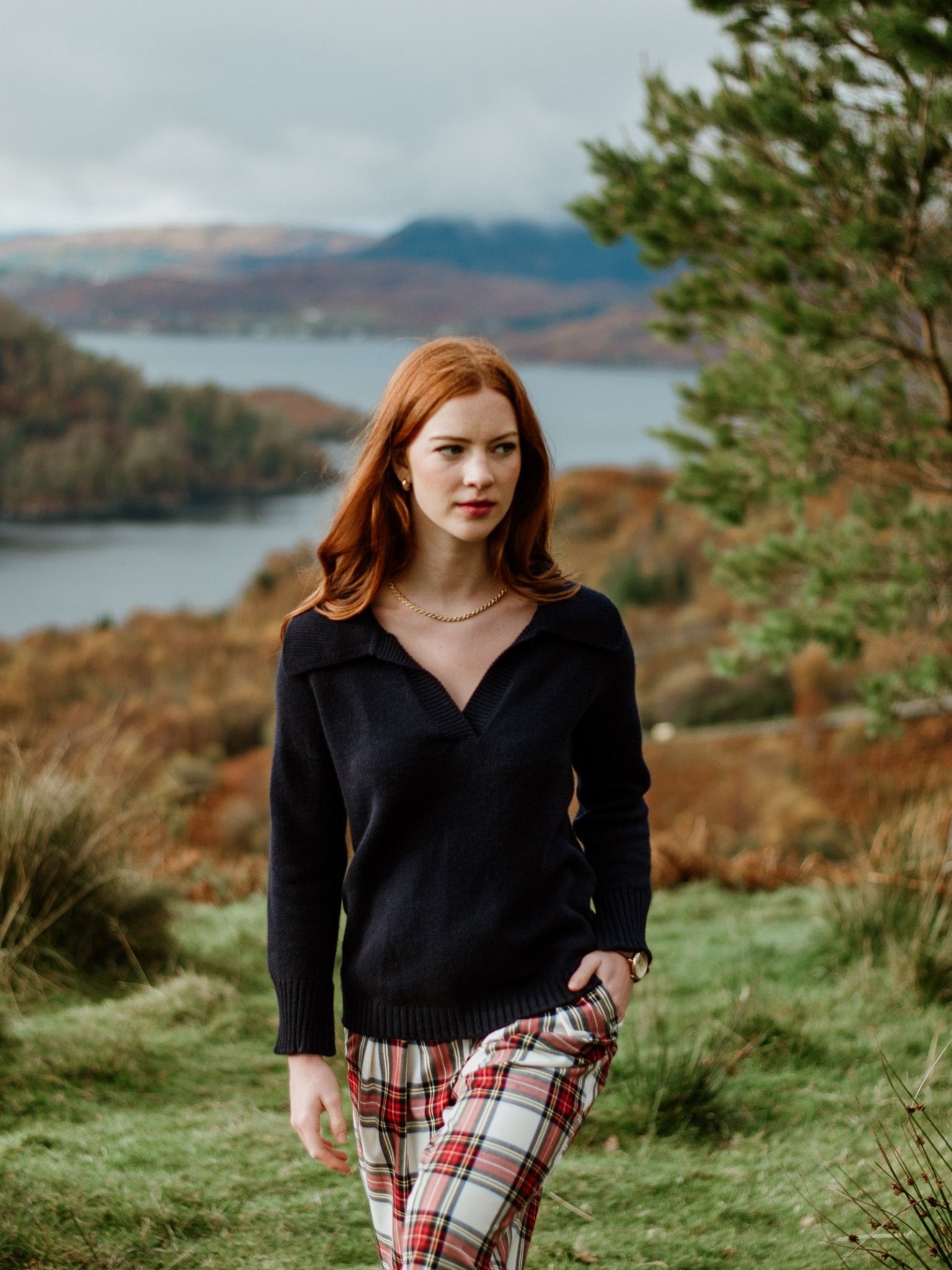 A woman with long red hair wears the Auld Stock Cashmere Shirt Jumper by Campbell's of Beauly and plaid pants, standing on grassy terrain with hills, trees, and a lake in the scenic, cloudy background.