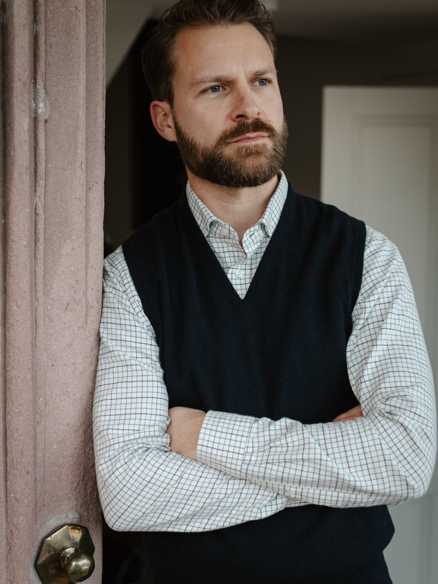 A bearded man stands indoors with arms crossed, wearing a Campbell's of Beauly Cashmere Slipover over a checkered shirt, leaning against a wall and gazing thoughtfully into the distance.