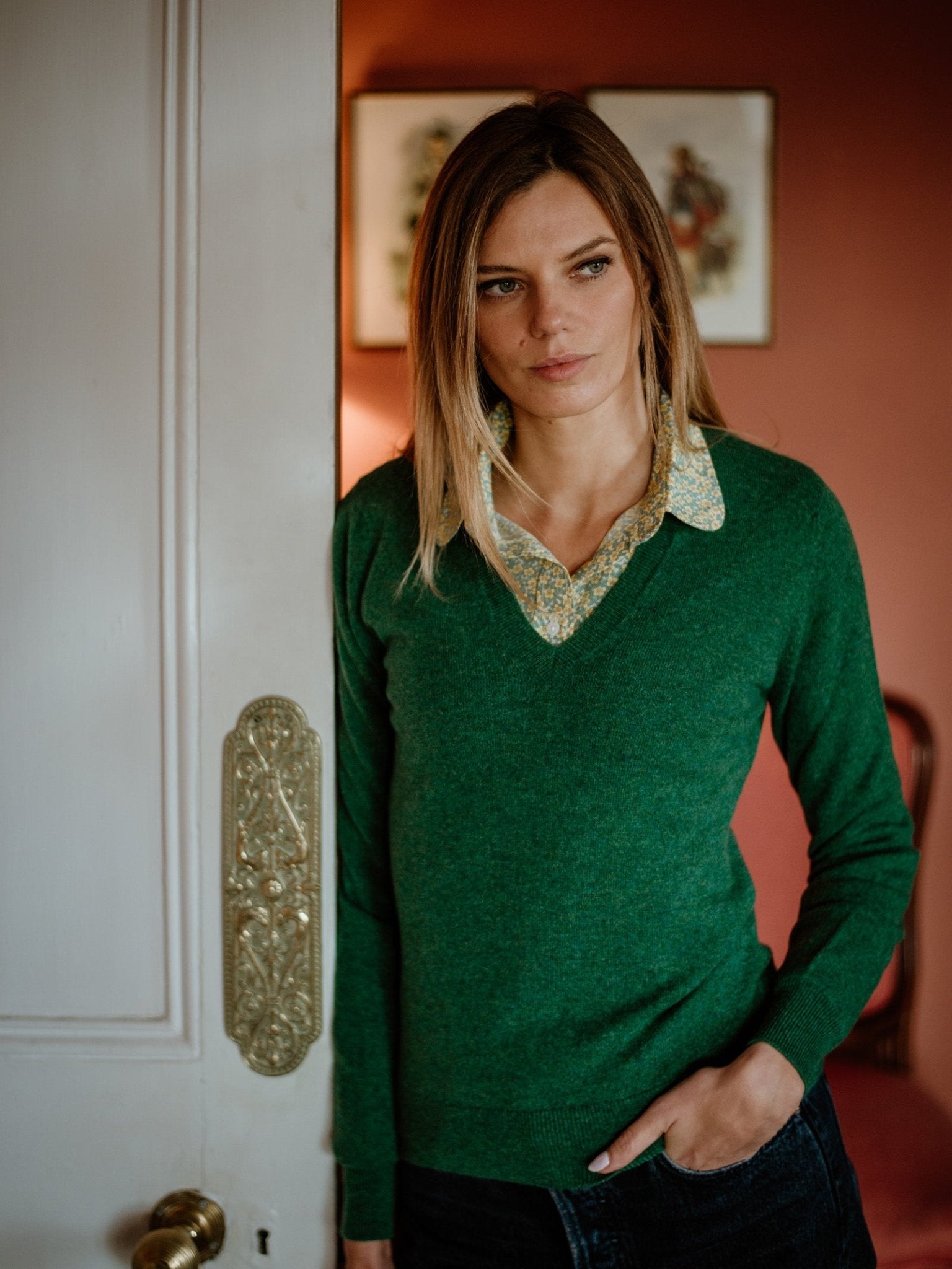 A woman with straight, light brown hair stands indoors by an open white door, wearing the Campbell's of Beauly Auld Stock Cashmere V-Neck Jumper over a collared shirt. She looks ahead thoughtfully; framed art decorates the warmly lit background.