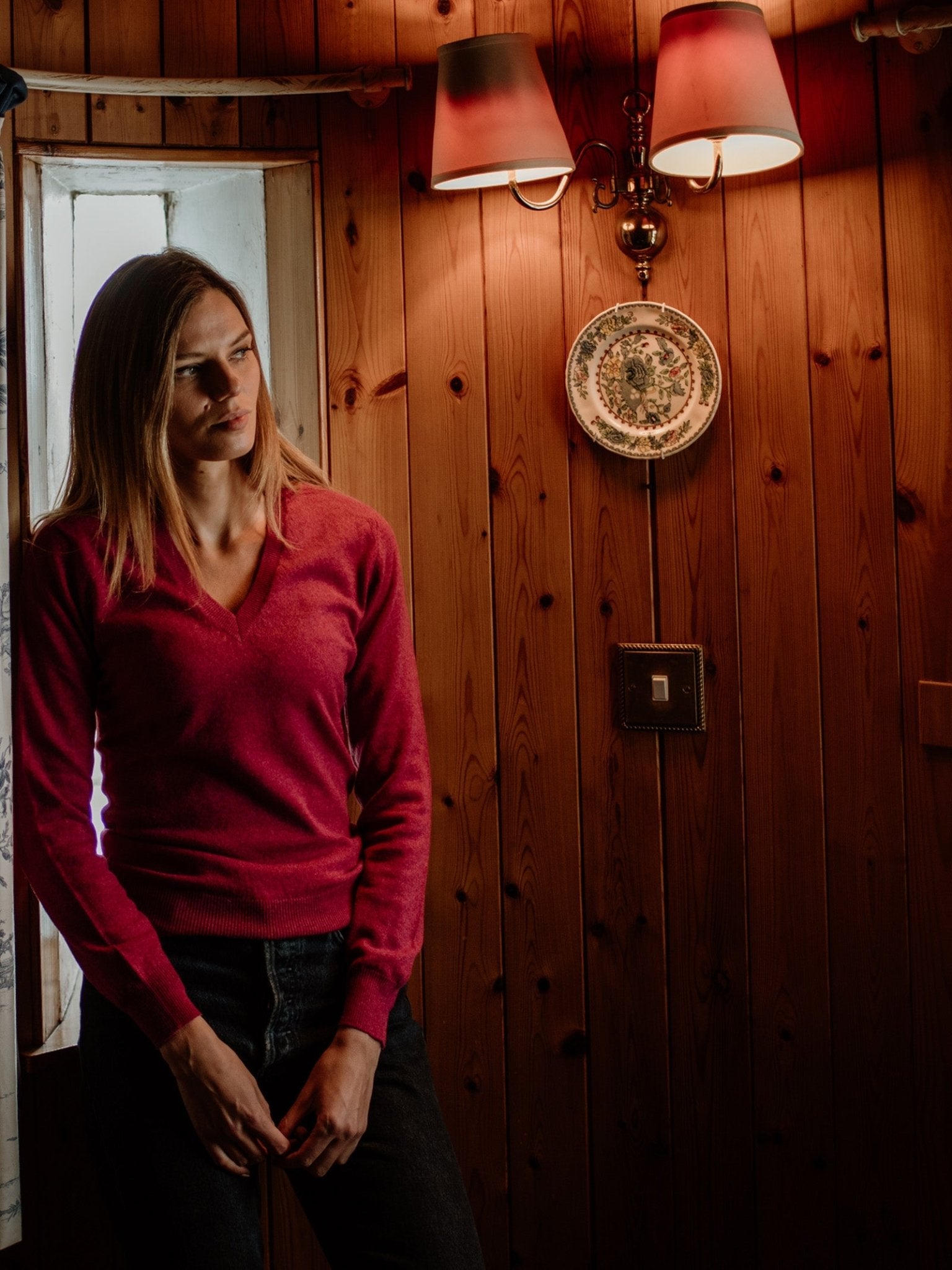 A person wearing the Campbell's of Beauly Auld Stock Cashmere V-Neck Jumper in red stands indoors by a wooden wall with décor and soft lighting, gazing thoughtfully toward the window in a cozy atmosphere.