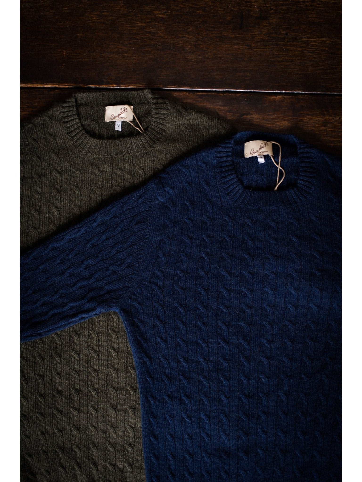 Two Campbell's of Beauly Cashmere Cable Crew Jumpers, one dark green and one navy, are laid flat on a dark wooden surface. Both feature ribbed trims and visible brand tags at the collar.
