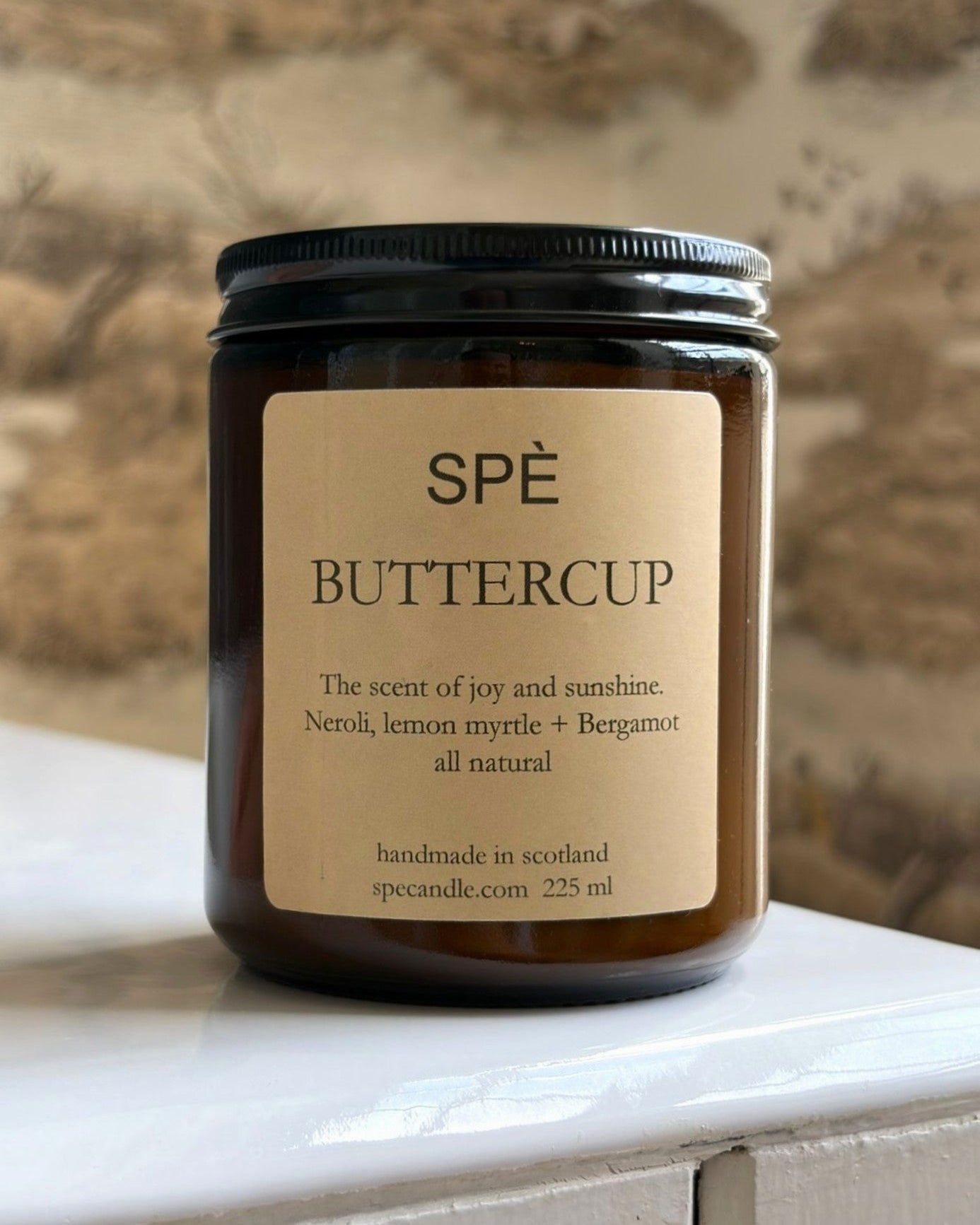 A brown candle jar labeled SPÈ Atelier Buttercup Candle, with scent notes of neroli, lemon myrtle, and bergamot, sits on a white surface against a blurred nature-inspired background, evoking the delicate fragrance of buttercups with an uplifting citrus floral aroma.