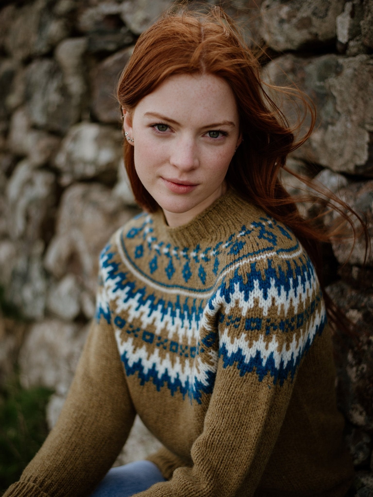 A young woman with long red hair wears the Auld Stock Chunky Fairisle Jumper by Campbell's of Beauly in brown, blue, and white Shetland wool, sitting calmly against a rustic stone wall and looking directly at the camera.