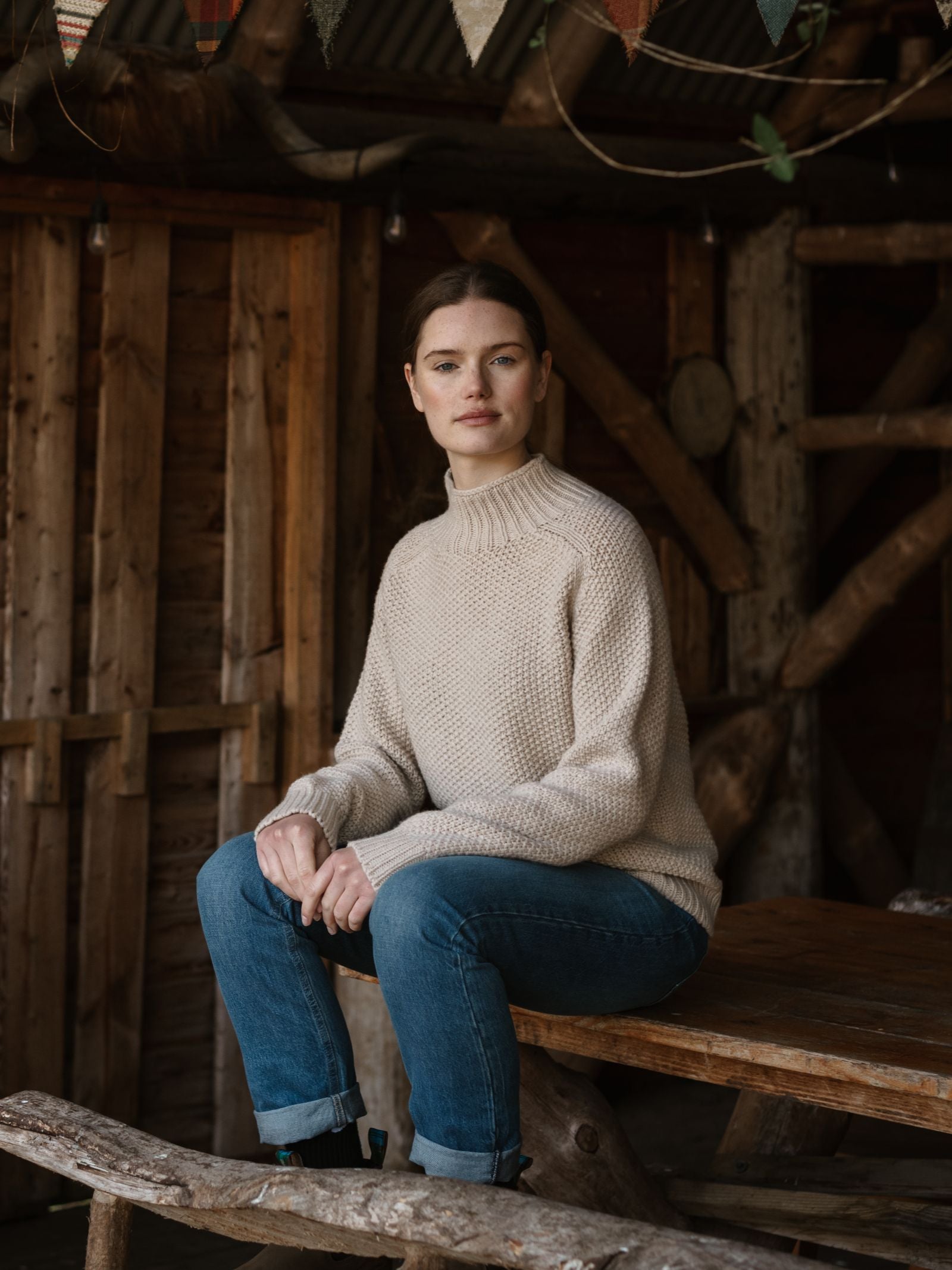A woman with light skin and brown hair tied back sits on a rustic wooden table in a cozy cabin, wearing Campbell's of Beauly's Chunky Moss Stitch Polo in oat and blue jeans. Bunting hangs above her as she looks calmly at the camera.