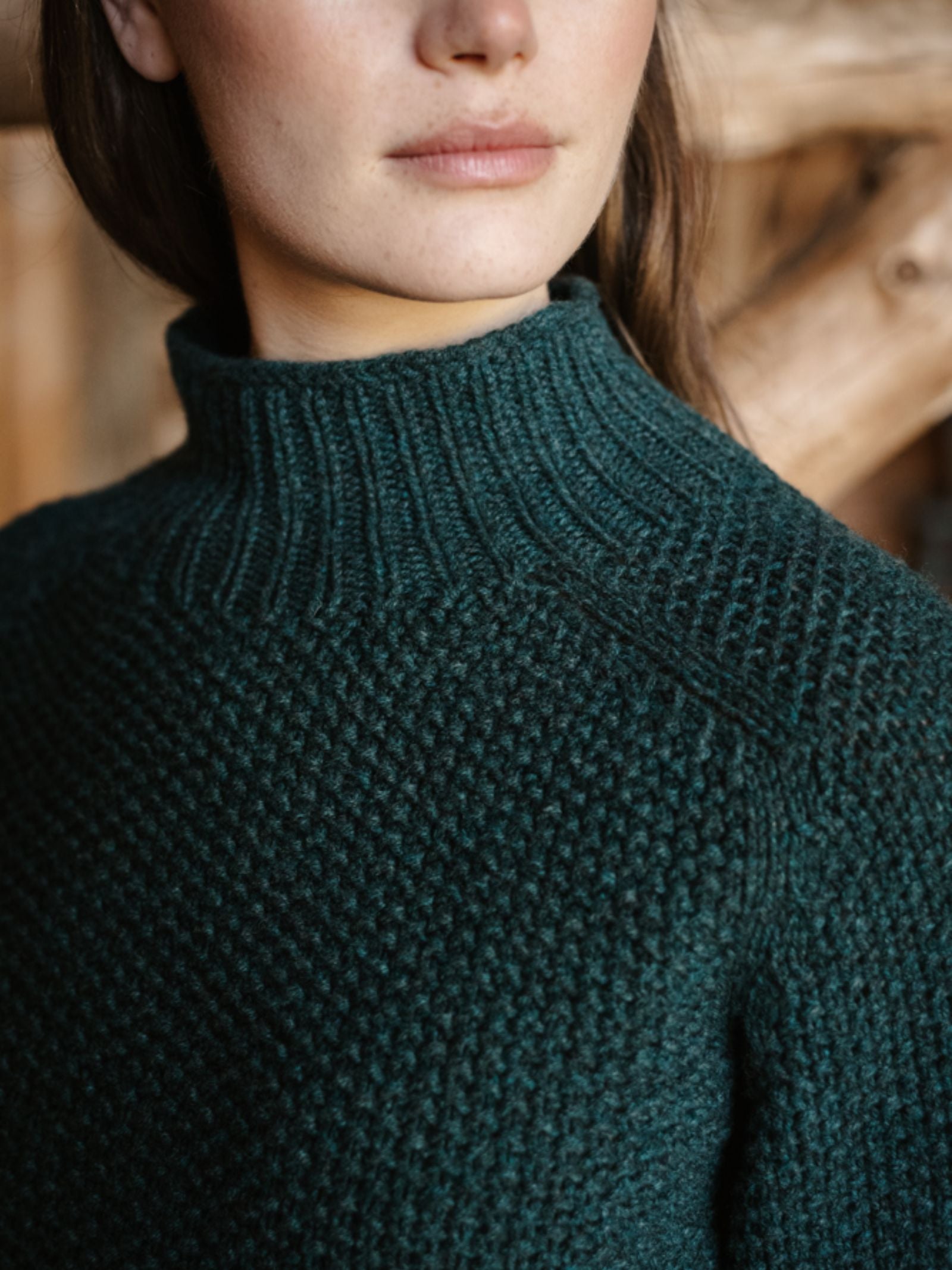 A close-up shows a person in the Chunky Moss Stitch Polo by Campbell's of Beauly, featuring a dark green chunky-knit lambswool high neck. Warm wooden tones fill the background, with the person's face partly visible.