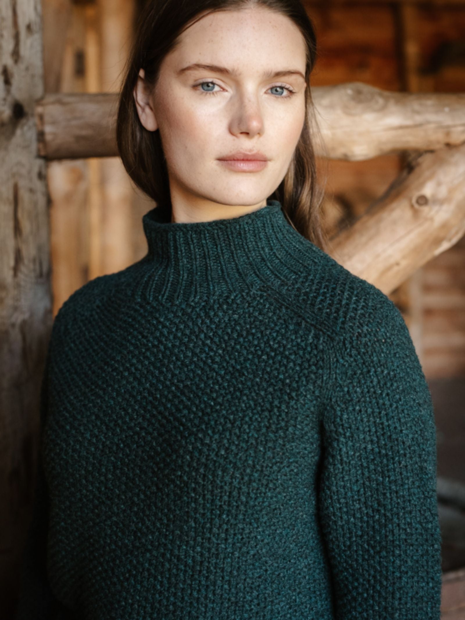 A woman with fair skin and long brown hair stands indoors, wearing the Campbell's of Beauly Chunky Moss Stitch Polo. Rustic wooden beams and a warmly lit interior complete the cozy setting.