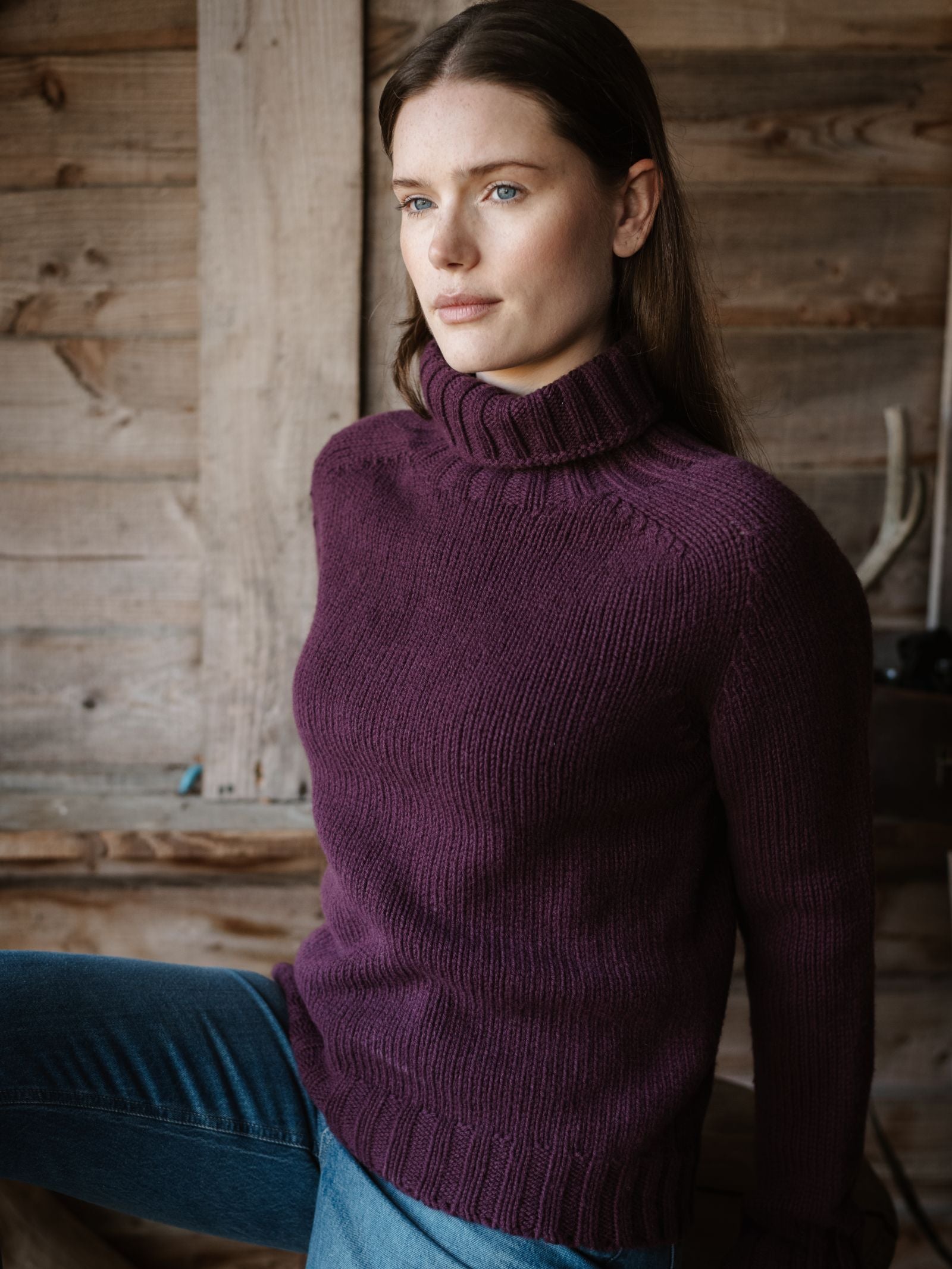 A woman with long brown hair and blue eyes, wearing a Campbell's of Beauly Chunky Roll Neck Sweater in purple and blue jeans, sits indoors by a rustic wooden wall, gazing thoughtfully into the distance.