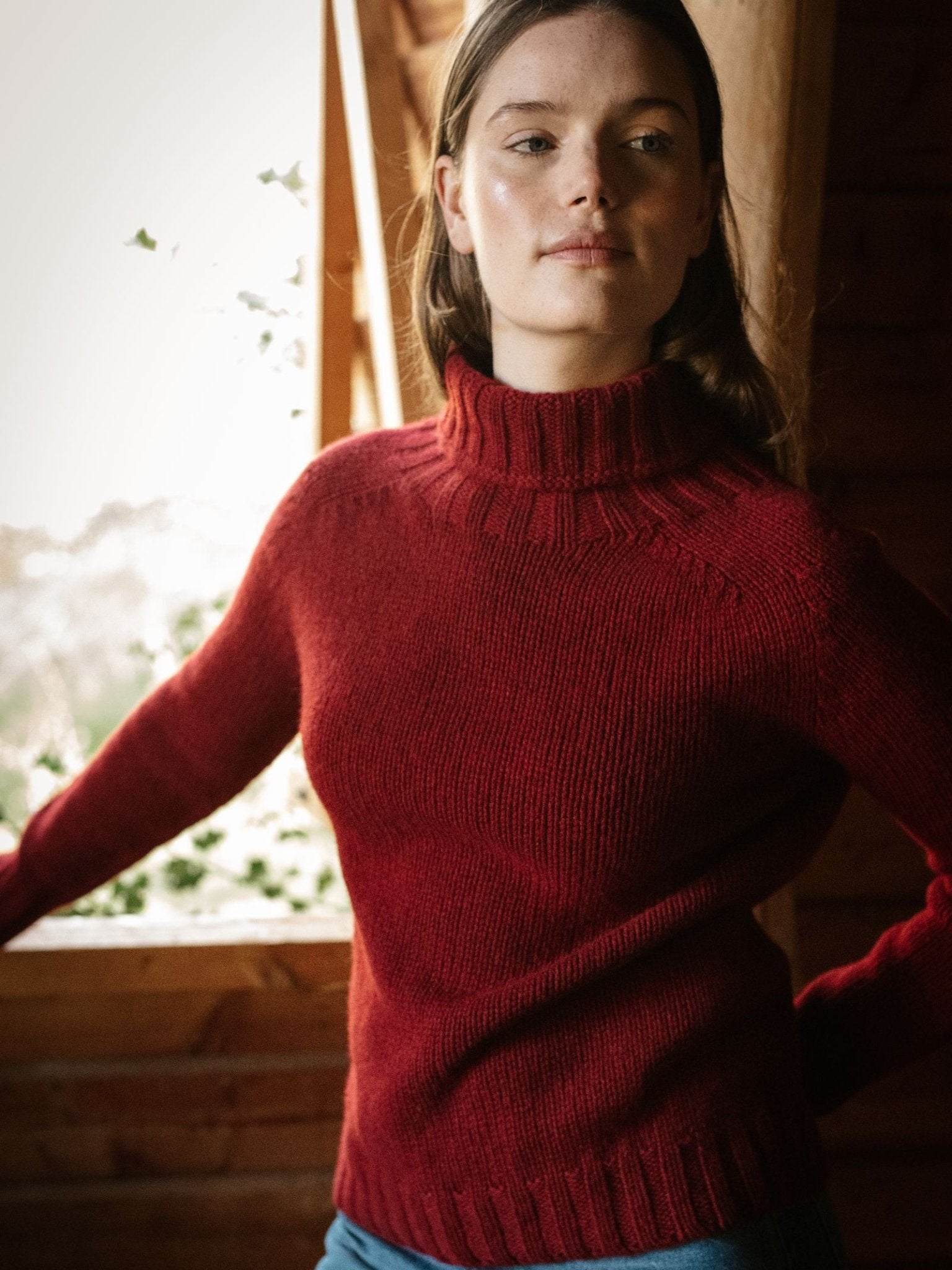 A woman poses indoors near a window, natural light on her face, wearing the Campbell's of Beauly Chunky Roll Neck Sweater in red. She stands before wooden walls, looking to the side with a relaxed expression.