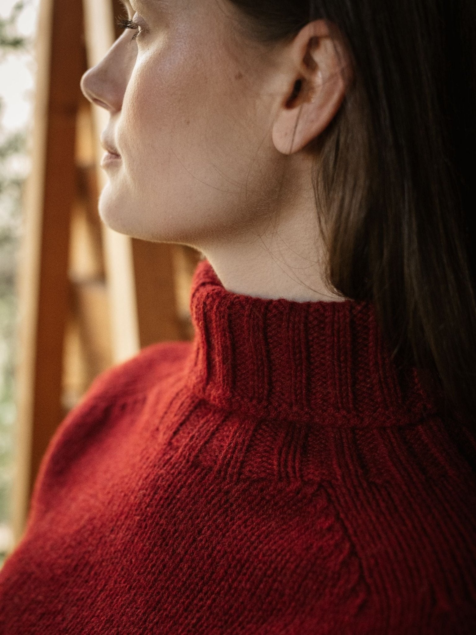 A close-up side view of a woman with long brown hair wearing the Campbell's of Beauly Chunky Roll Neck Sweater, a supersoft wool red turtleneck, standing by a wooden structure with soft natural light on her face.
