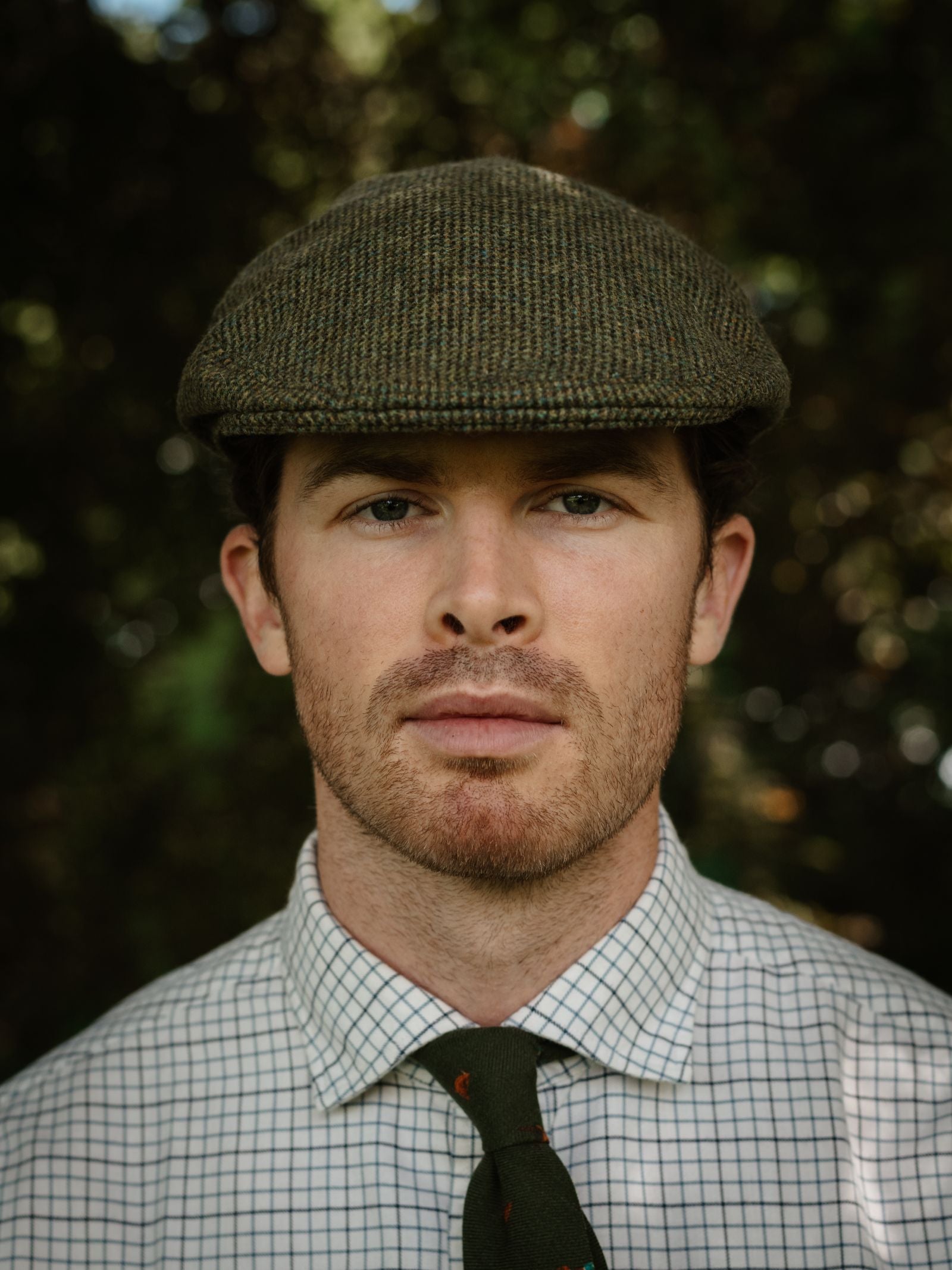 A person wearing the Campbell's of Beauly Unisex Green Conon Cap, a white checkered shirt, and a green tie stands outdoors before leafy greenery, looking directly at the camera with a neutral expression.