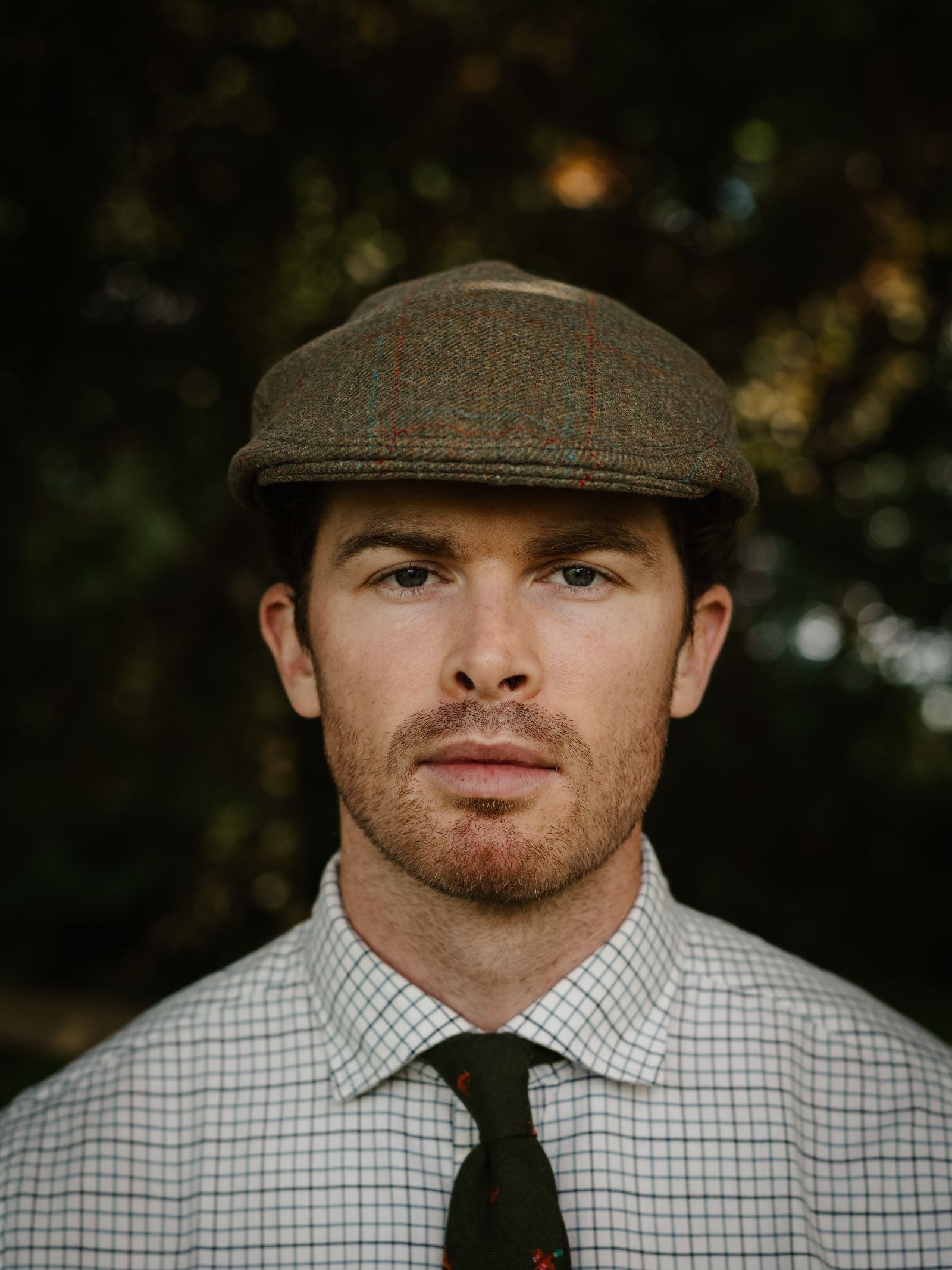 A man with fair skin and short brown hair wears the Campbell's of Beauly Unisex Brown Conon Cap, a checkered shirt, and a dark green tie. He looks directly at the camera with a neutral expression against a blurred green background.