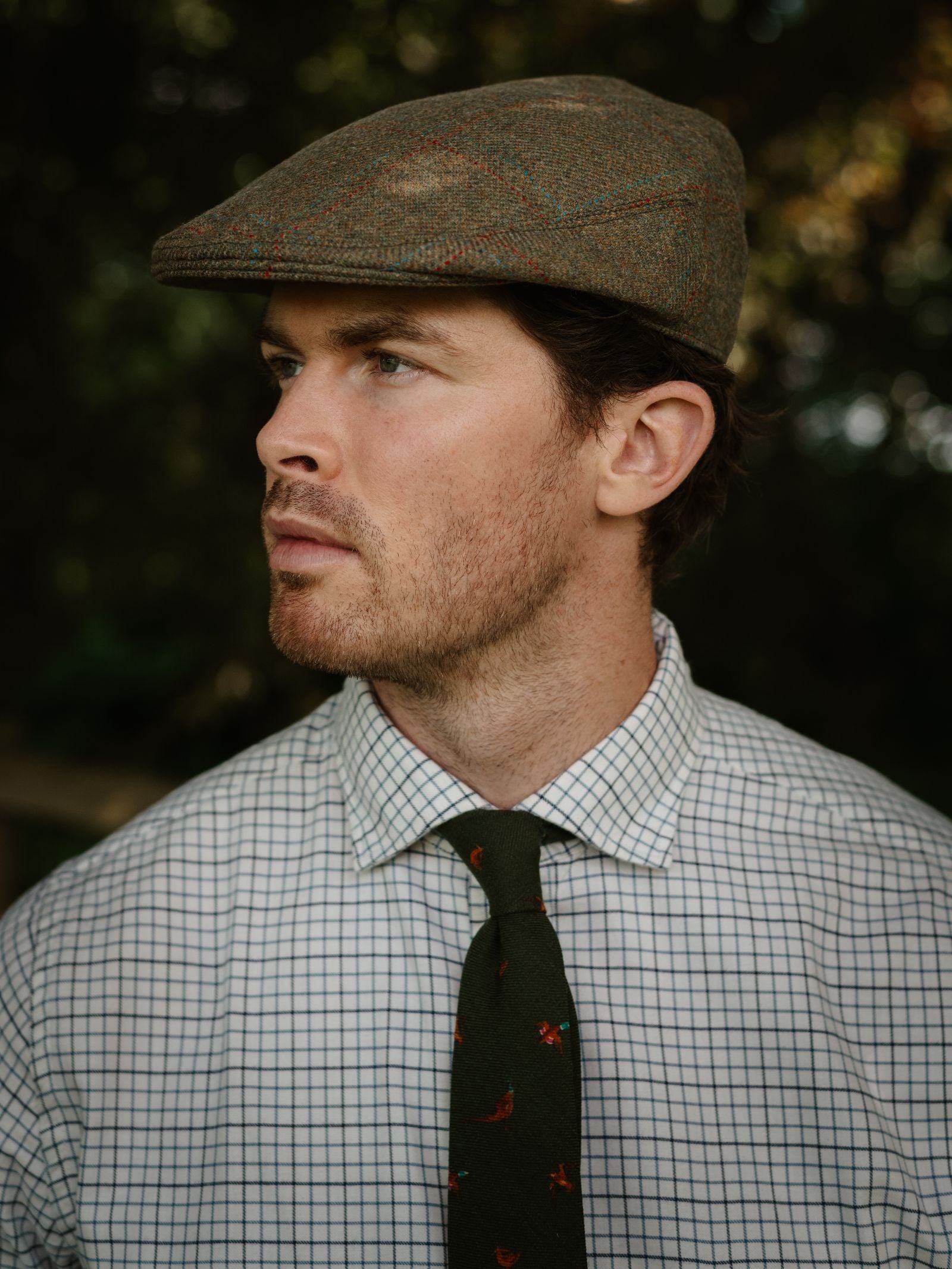 A man in a Campbell's of Beauly Unisex Brown Conon Cap, a white checkered shirt, and a green tie with small red designs looks to the side against a blurred outdoor backdrop.