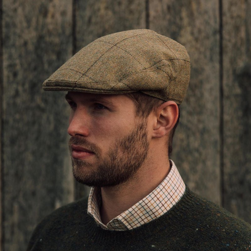 A man with a short beard wears the Campbell's of Beauly Auld Stock Conon Cap, along with a checked collared shirt and dark green sweater, standing before a rustic wooden wall.