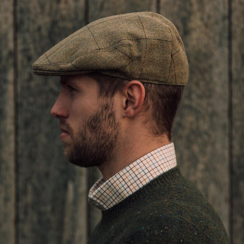 A bearded man in profile wears the Campbell's of Beauly Auld Stock Conon Cap, a green sweater, and a checked shirt, standing before a wooden textured background.