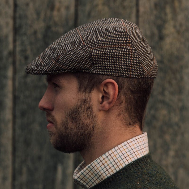 A man with a short beard, dressed in a green sweater and checkered shirt, stands in profile against a rustic wooden backdrop, wearing the Unisex Brown Conon Cap by Campbell's of Beauly.