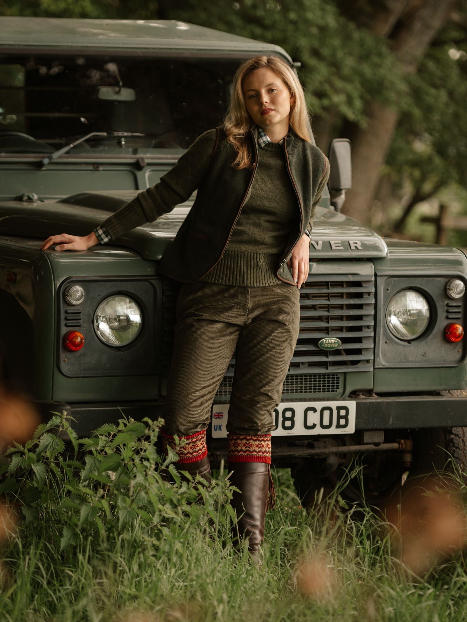Wearing Campbell's of Beauly Corduroy Plus Twos and brown boots, a woman in classic sporting tweeds leans against a green Land Rover on grassy ground, with trees visible in the background.