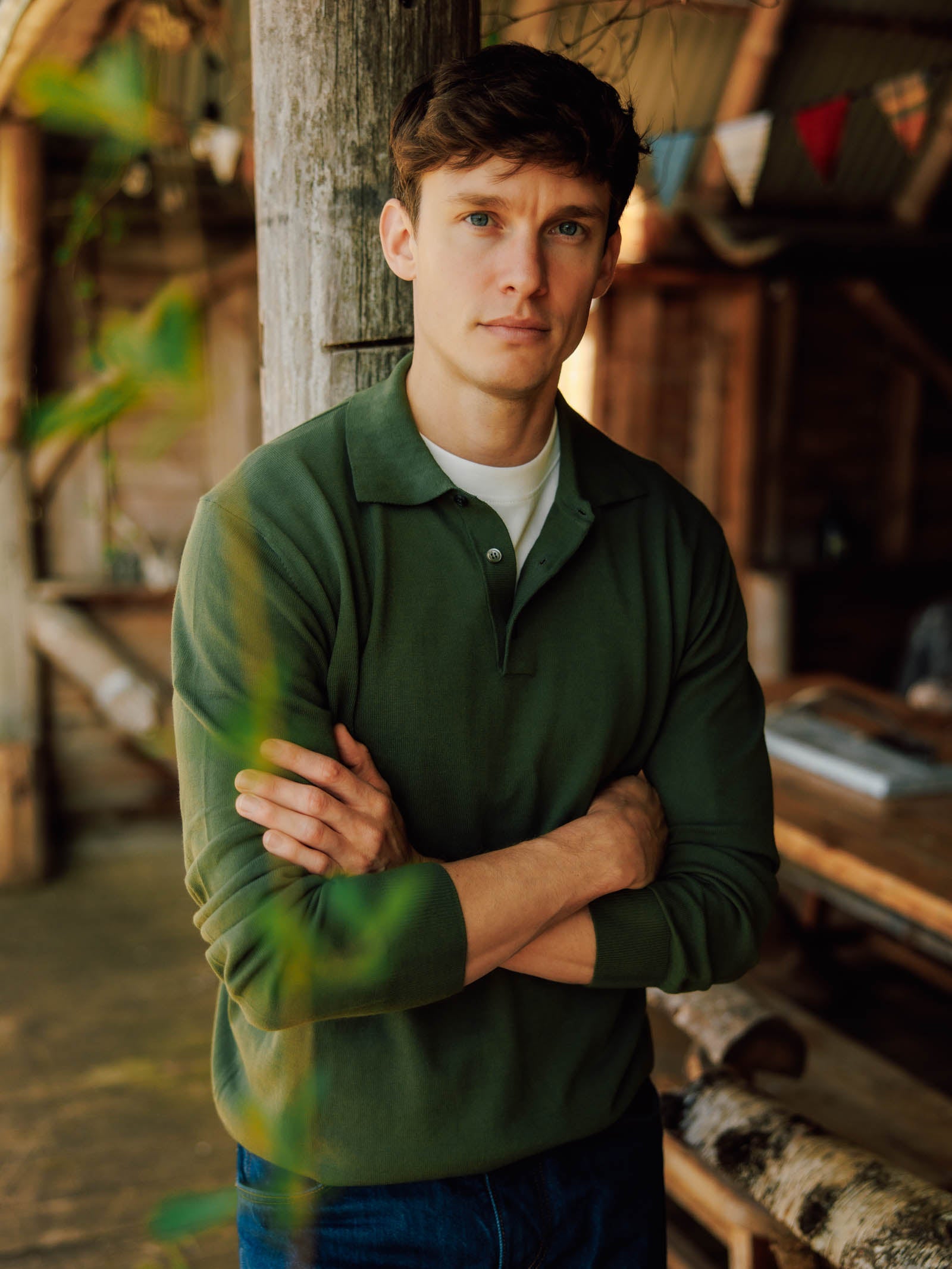 A man with short brown hair stands indoors, arms crossed and leaning against a wooden post, wearing the Campbell's of Beauly Cotton Rugby. Festive bunting and rustic wooden furniture are in the background.