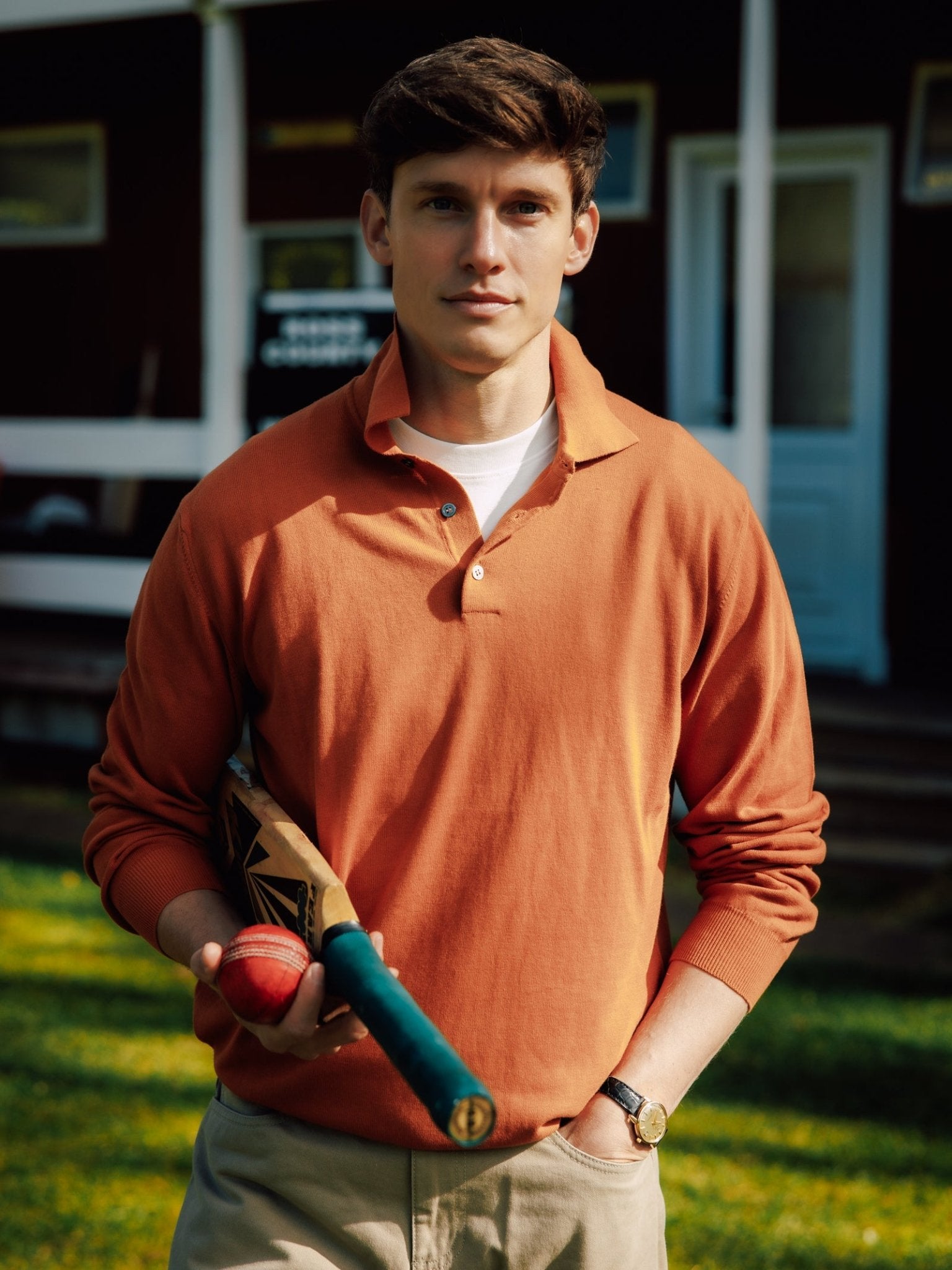 A young man outdoors holds a cricket bat and ball, dressed in a Campbell's of Beauly Cotton Rugby and khaki pants, with a building and green lawn in the background.